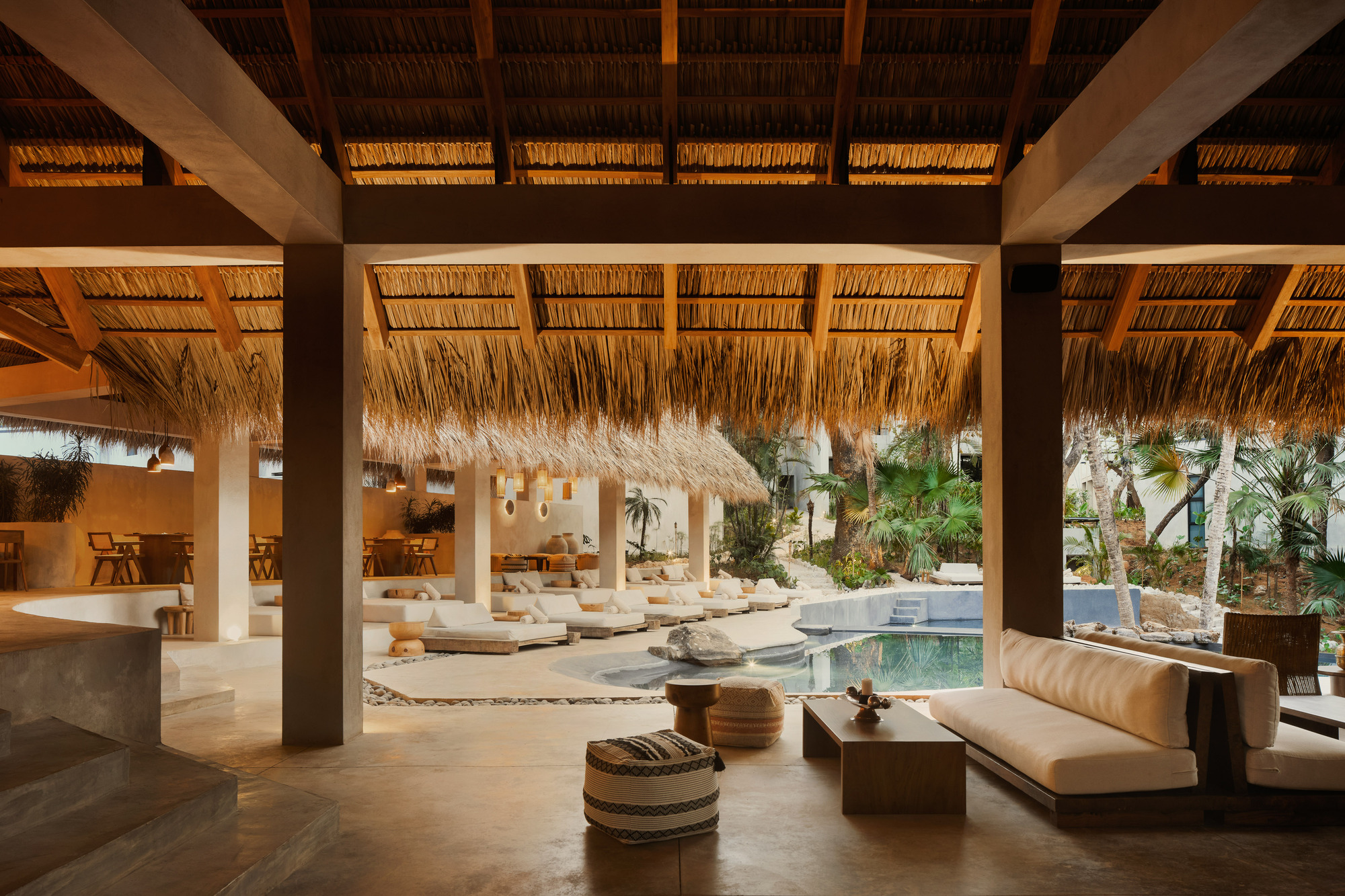 Open lounge under a palm-thatch roof on timber beams, with daybeds, a pool and palms beyond in warm evening light