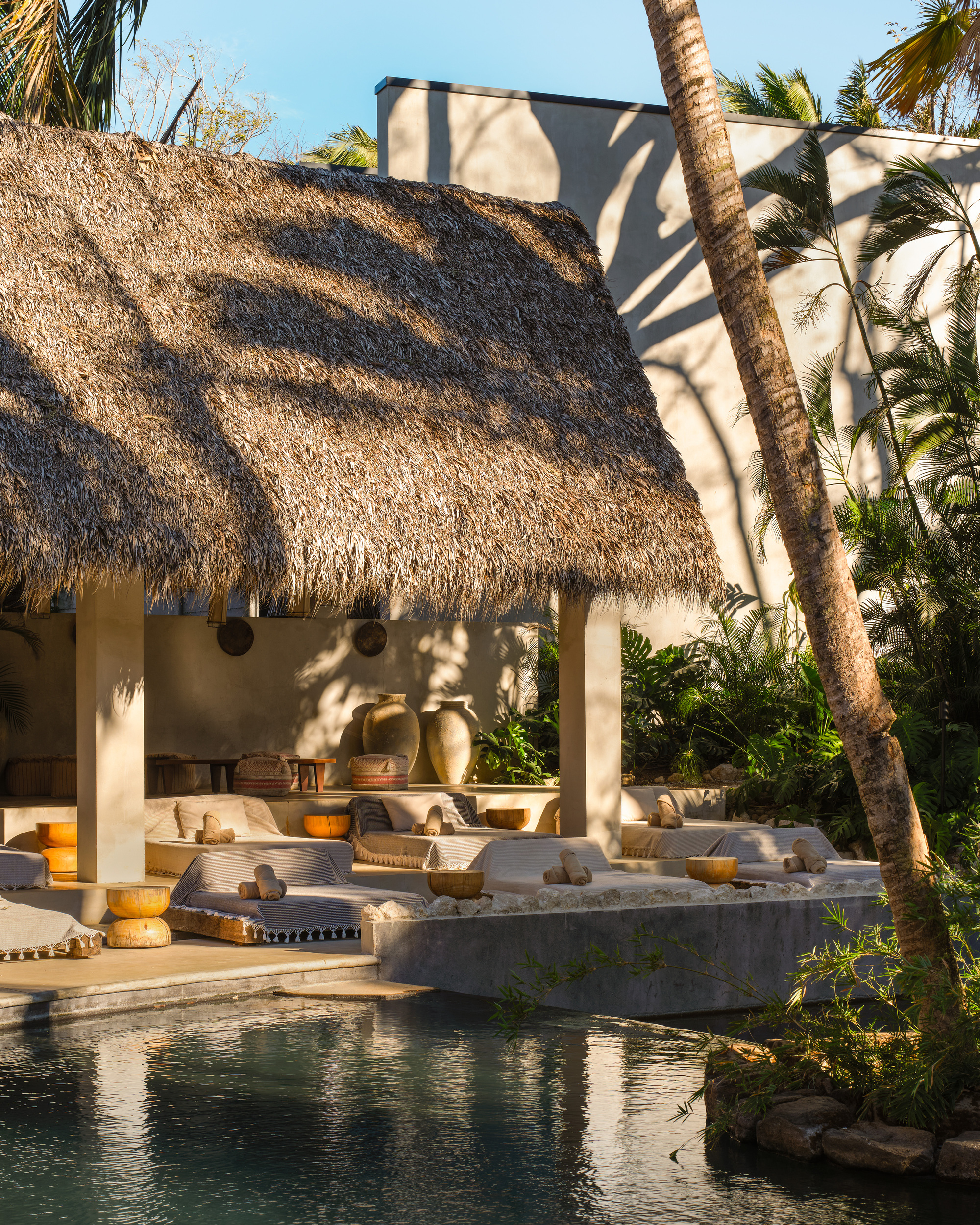 Thatched pavilion with raised daybeds and clay vessels beside a pool, palm shadows falling across the pale plaster wall