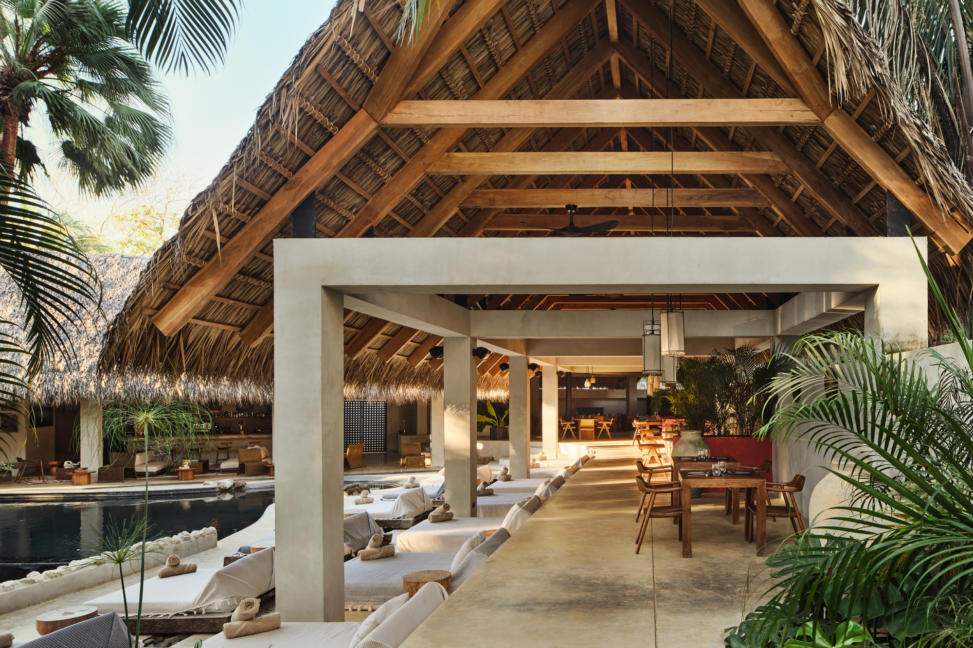 Concrete portal frame opening onto a thatched timber-trussed pavilion, with daybeds along the pool edge and a dining table beyond