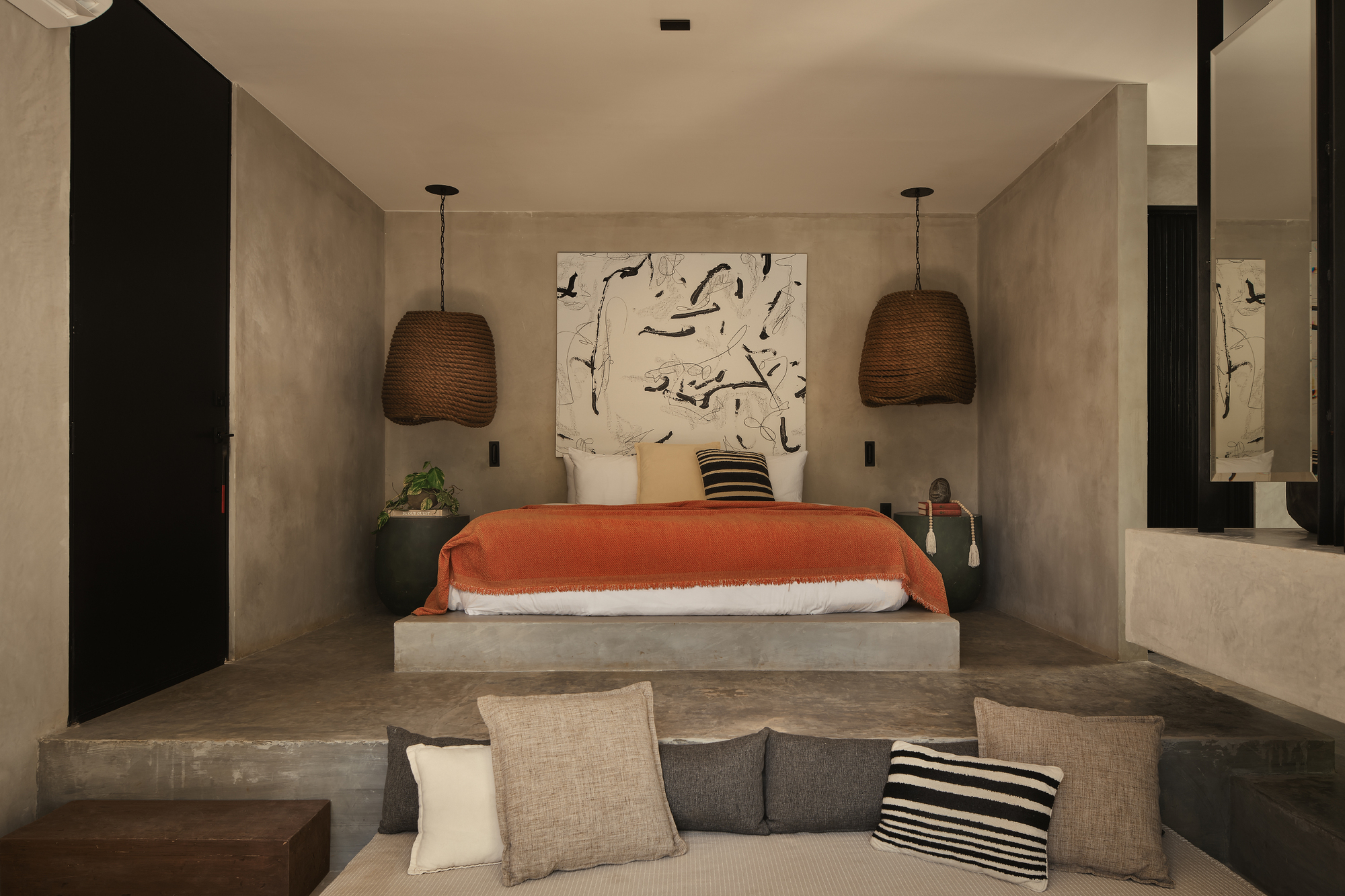 Bedroom with a raised concrete platform bed, orange throw, woven pendant lamps and abstract artwork above a cushioned seat