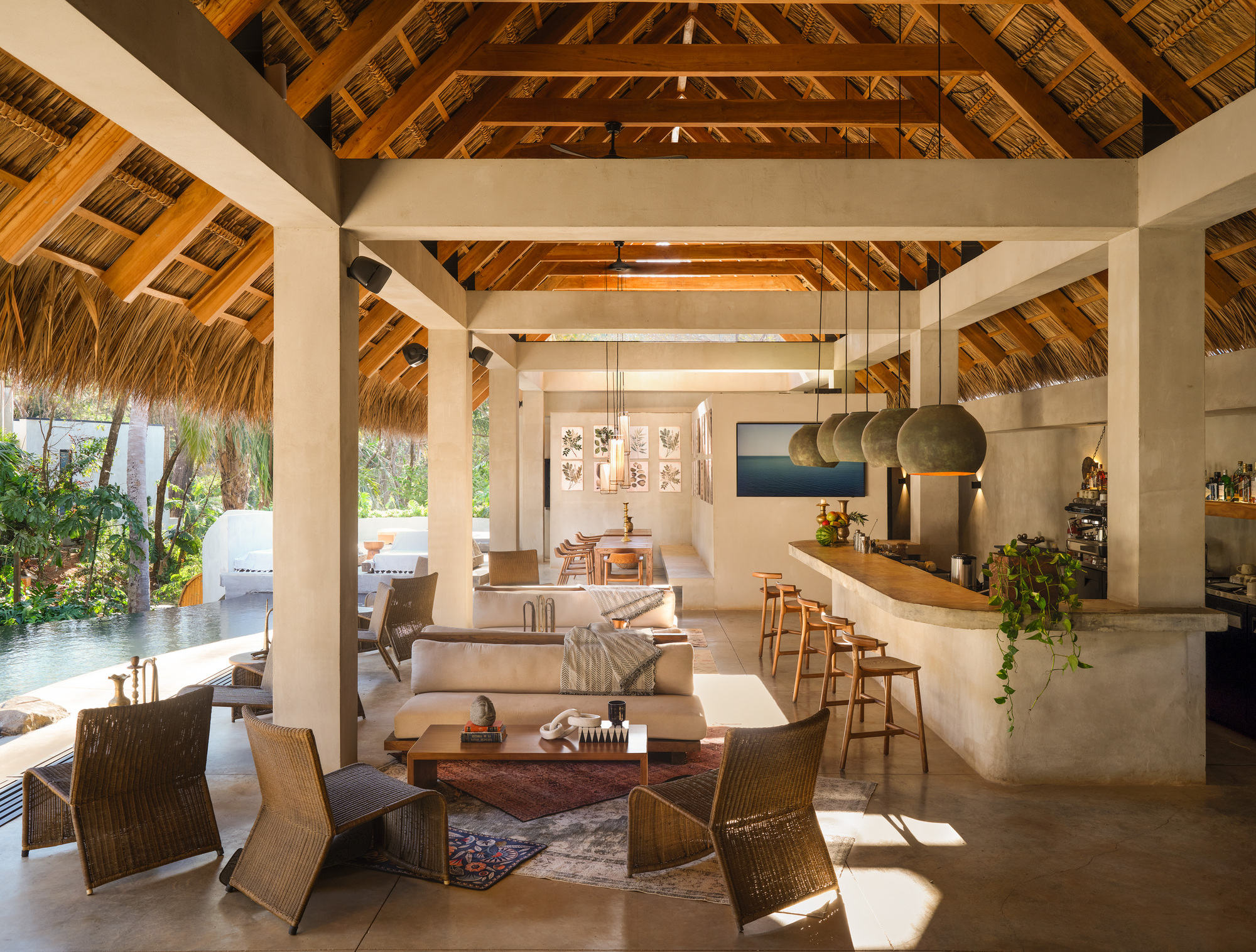 Open lounge under a thatched timber roof with woven chairs, clay pendant lamps and a curved concrete bar beside the pool in afternoon sun