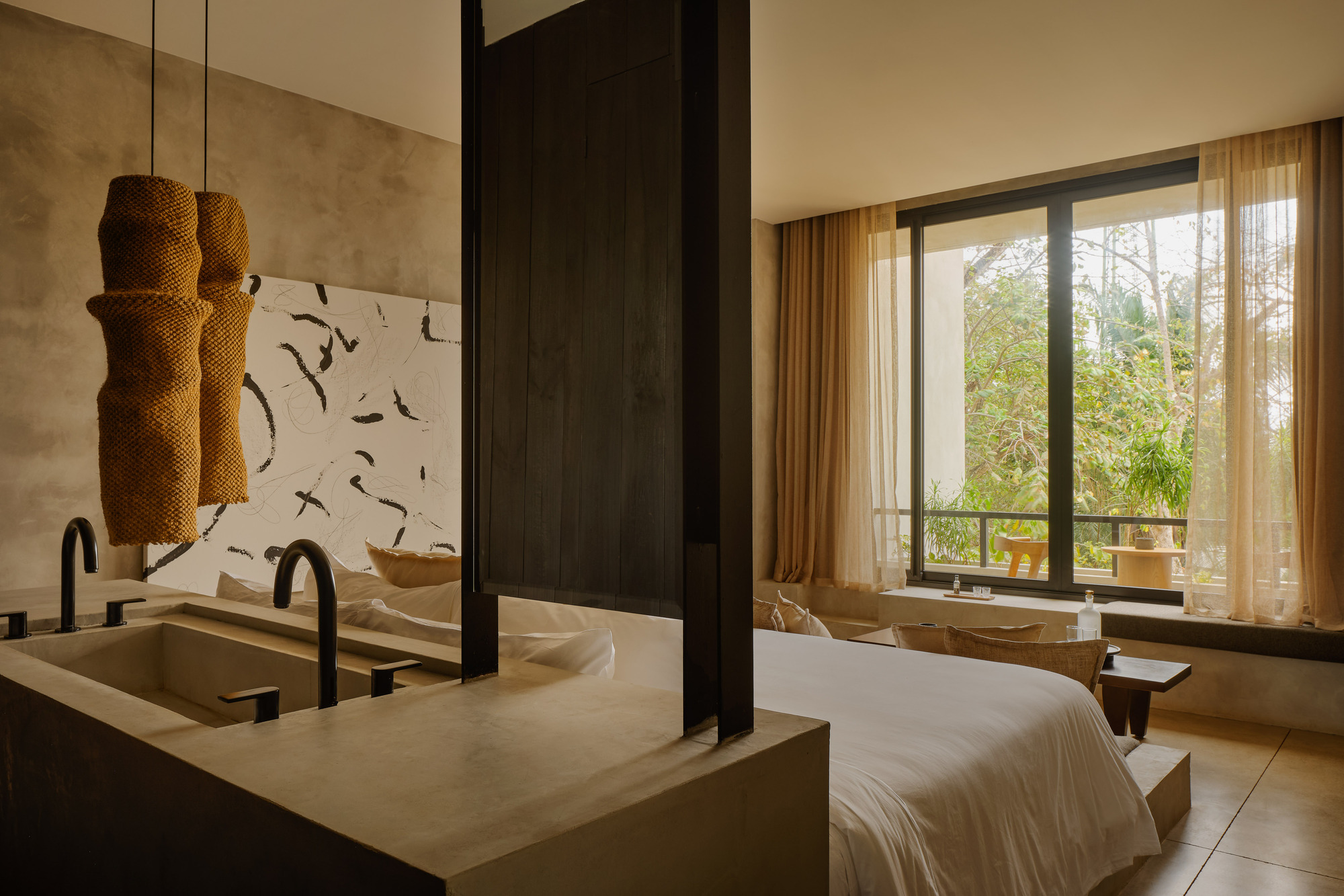 Bedroom with a concrete basin and black taps, woven pendant lamps and sheer curtains framing a tall window onto trees