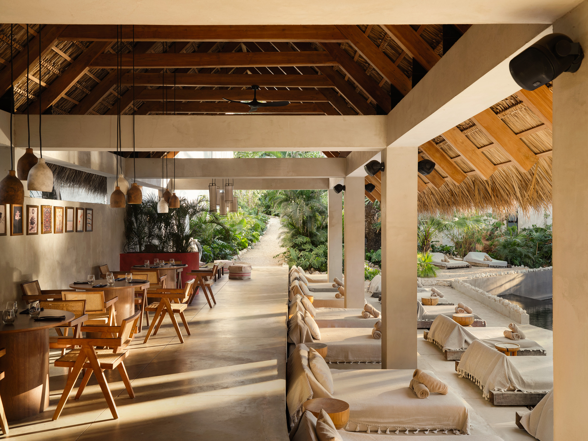 Covered walkway with timber dining chairs on one side and daybeds on the other, thatched roof overhead and morning light on the floor
