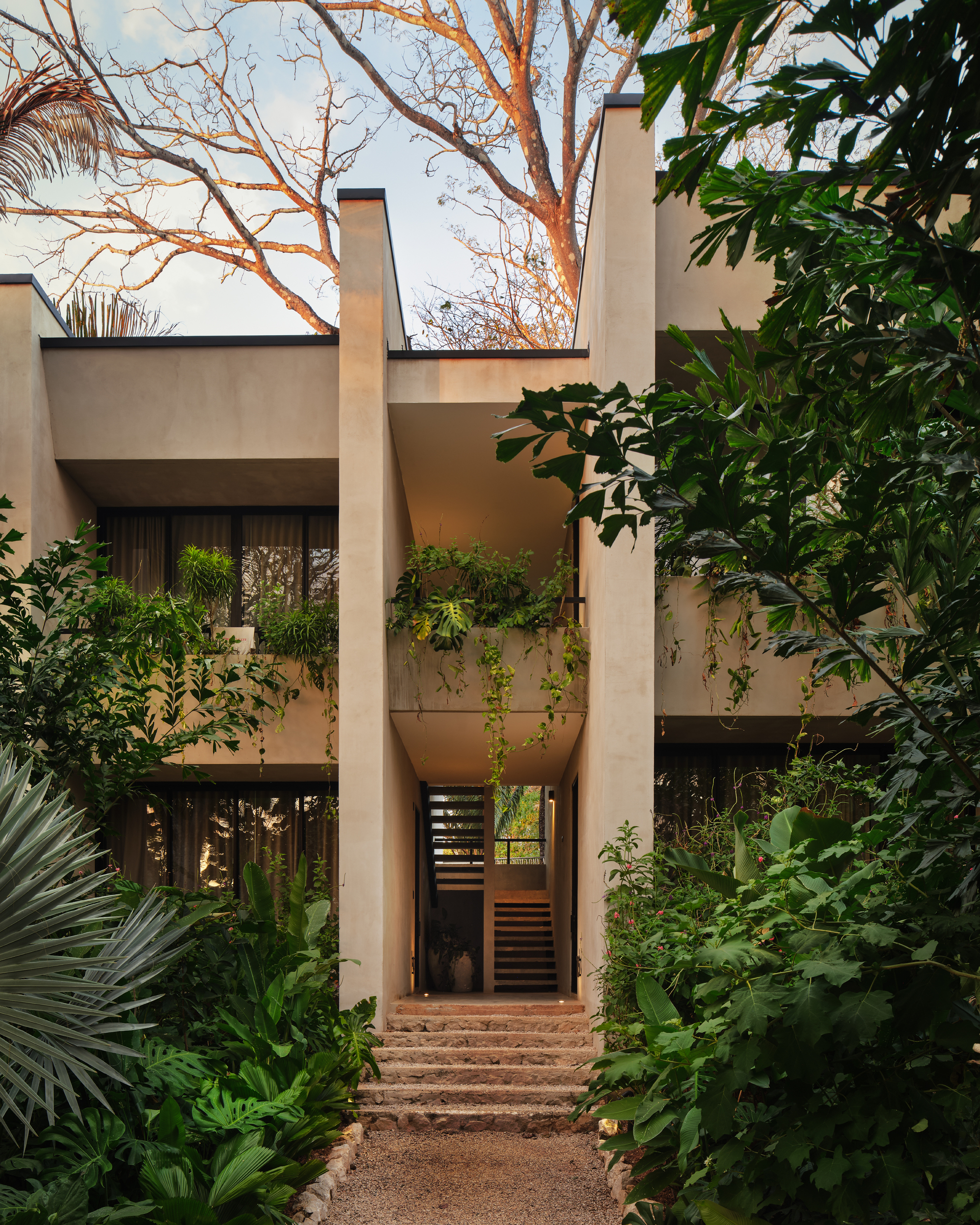 Two-storey stucco facade with planted balconies and a stone stair rising between tall piers, under bare branches at dusk