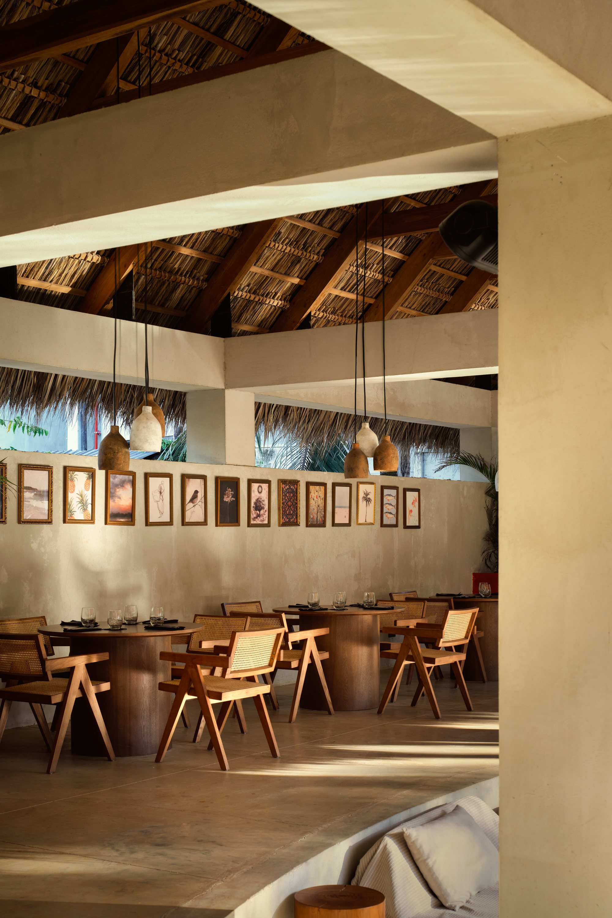 Dining room with round wooden tables, cane chairs and pendant lamps beneath a thatched roof, with framed botanical prints on the wall