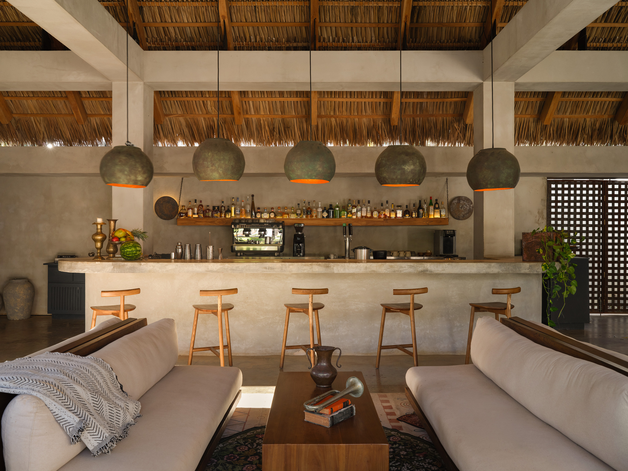 Open-air bar with a concrete counter, wooden stools and patinated pendant lamps beneath a thatched palm roof, glowing in warm evening light
