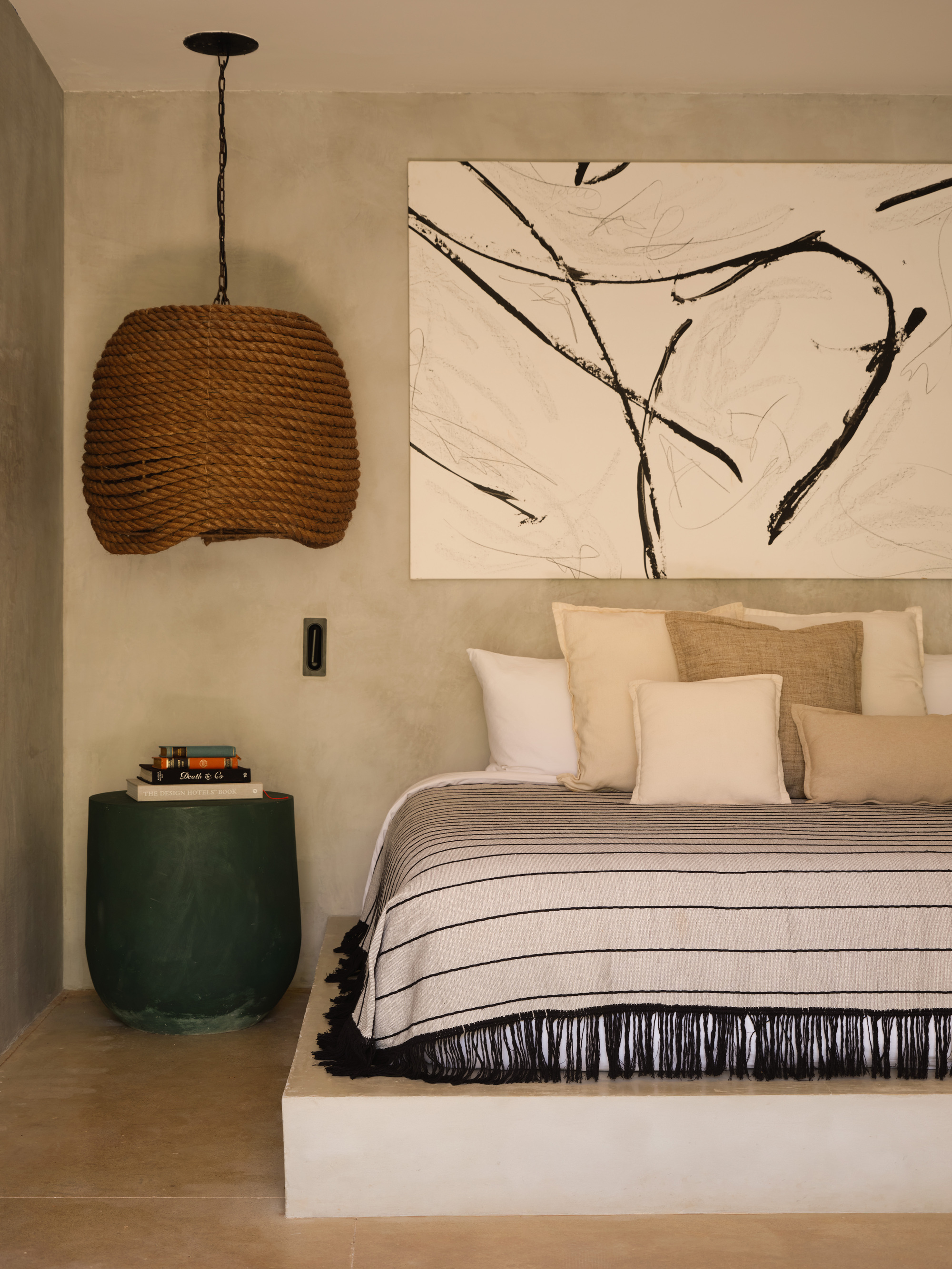 Rope pendant lamp beside a green side table and a striped fringed bedcover, with a black ink painting above the pillows
