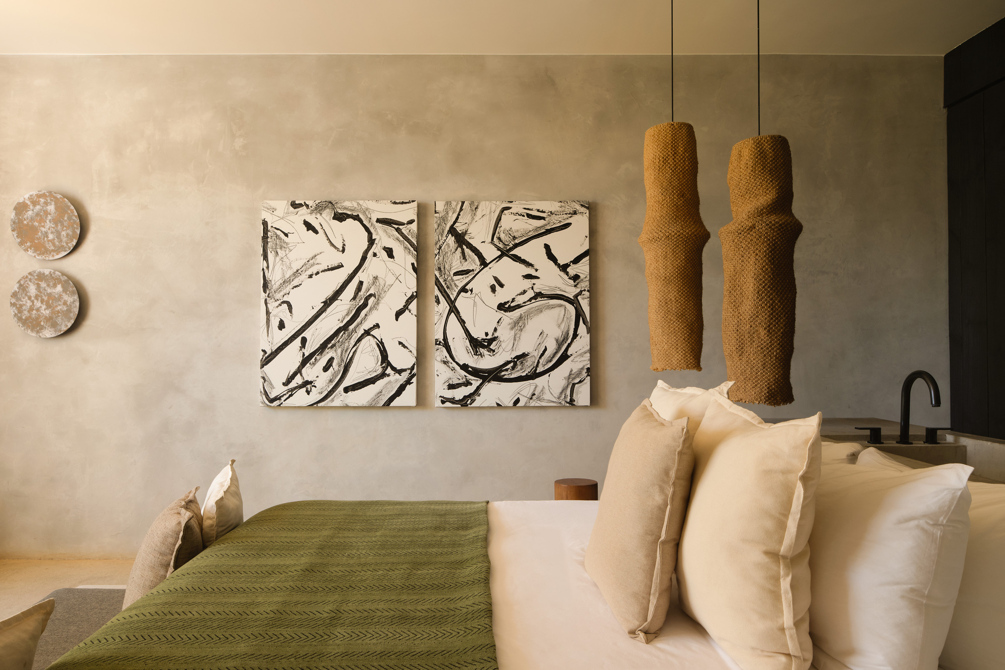 Two brushstroke canvases and woven cylindrical pendants above an olive green bed throw, with a black faucet at the right edge