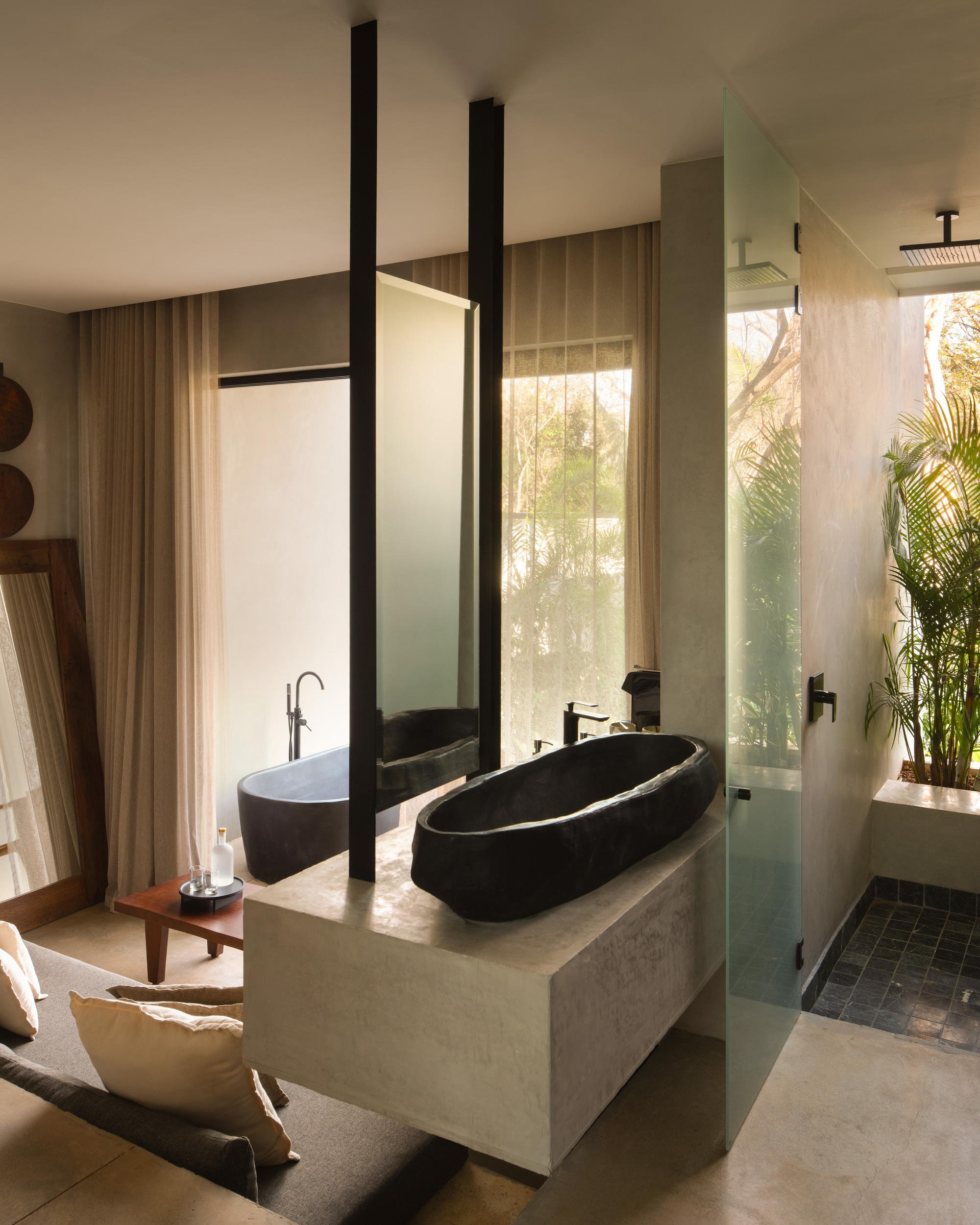 Carved black stone basin on a plaster vanity, with a freestanding tub, sheer curtains and palms seen through the frosted glass shower door