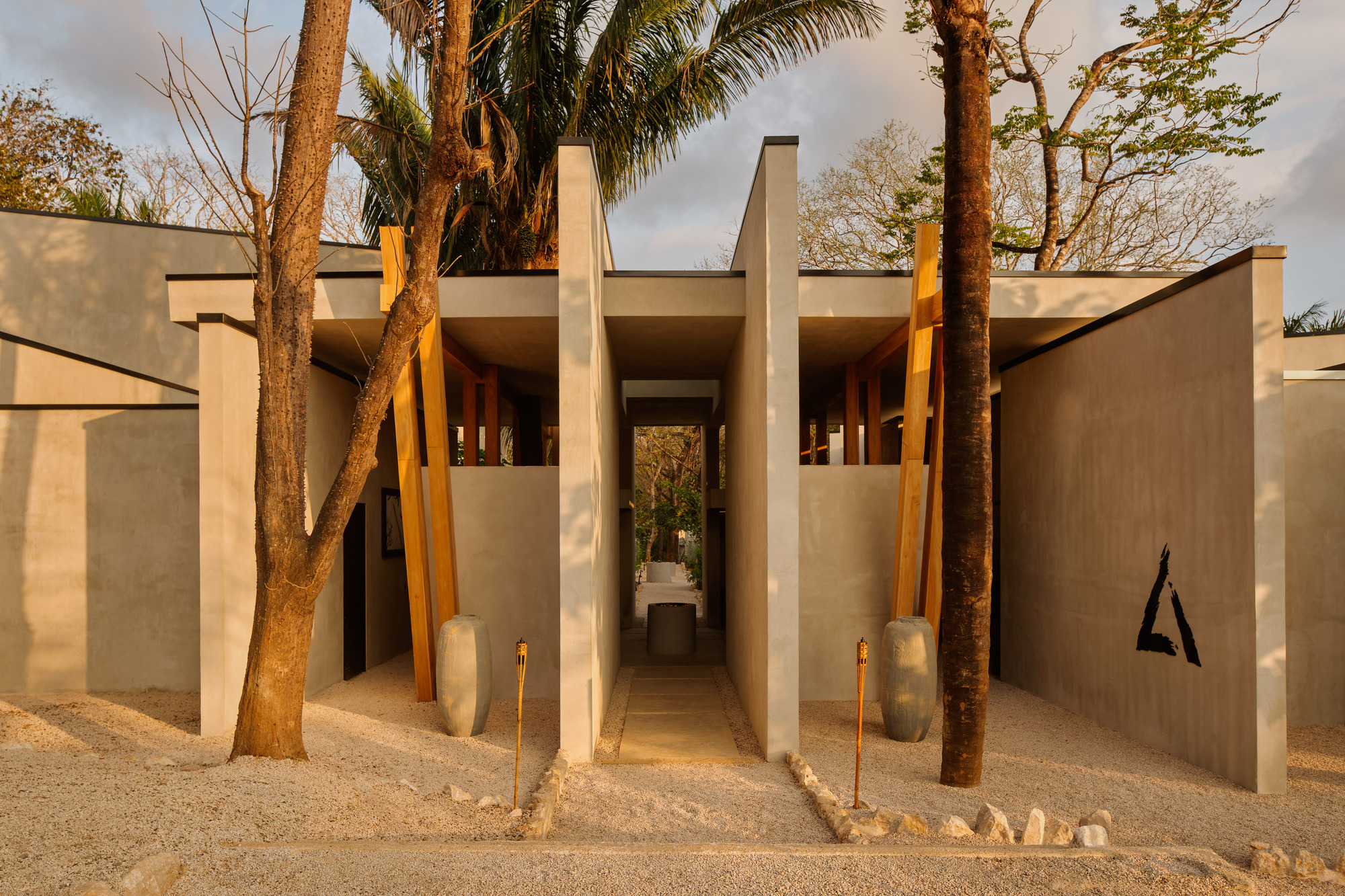 Concrete entry passage with slanted timber struts and clay jars, lit by low evening sun through bare trees