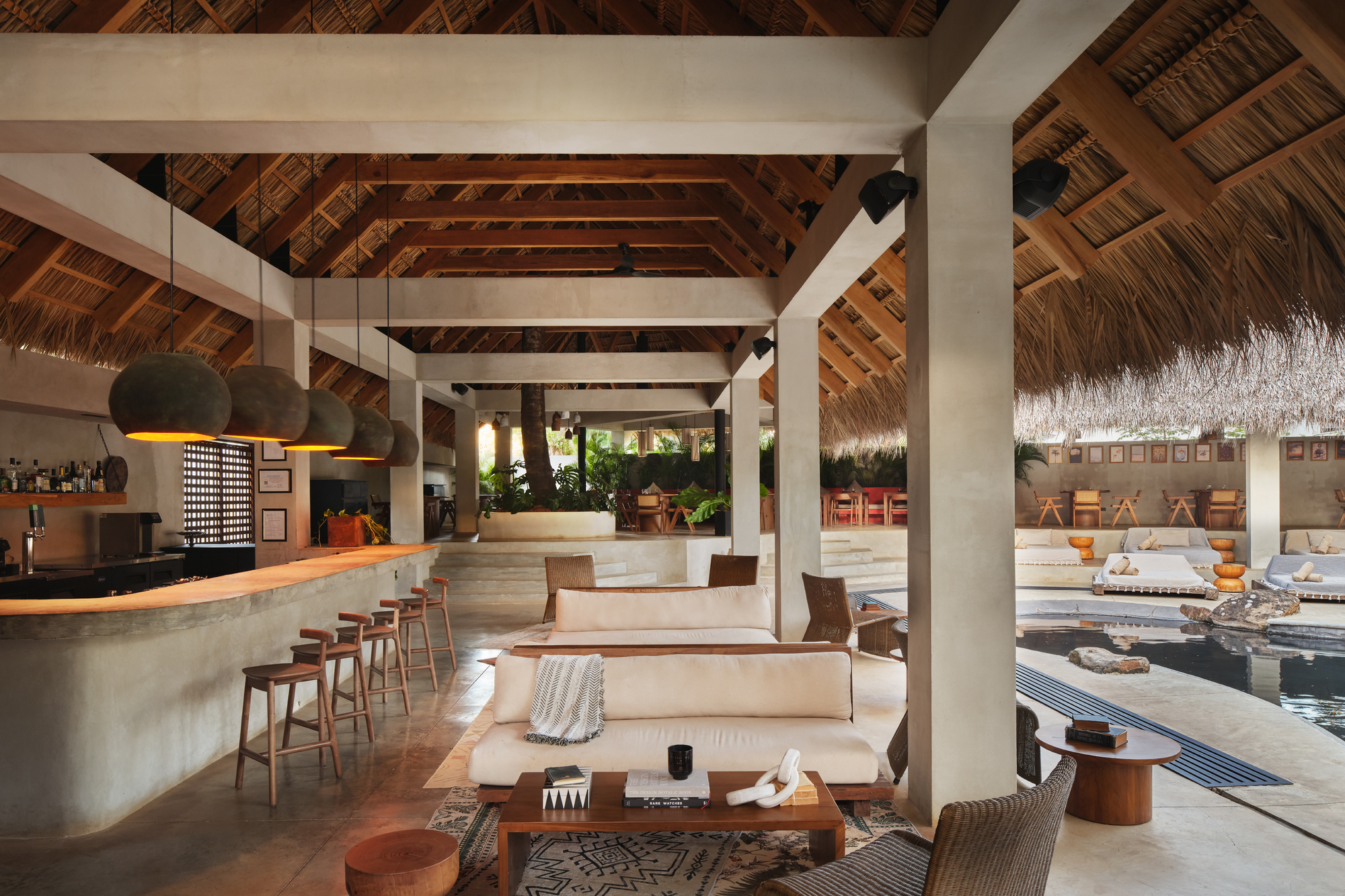 Open lounge under a thatched timber roof, with a curved bar, pendant lamps and a pool beyond the sofas