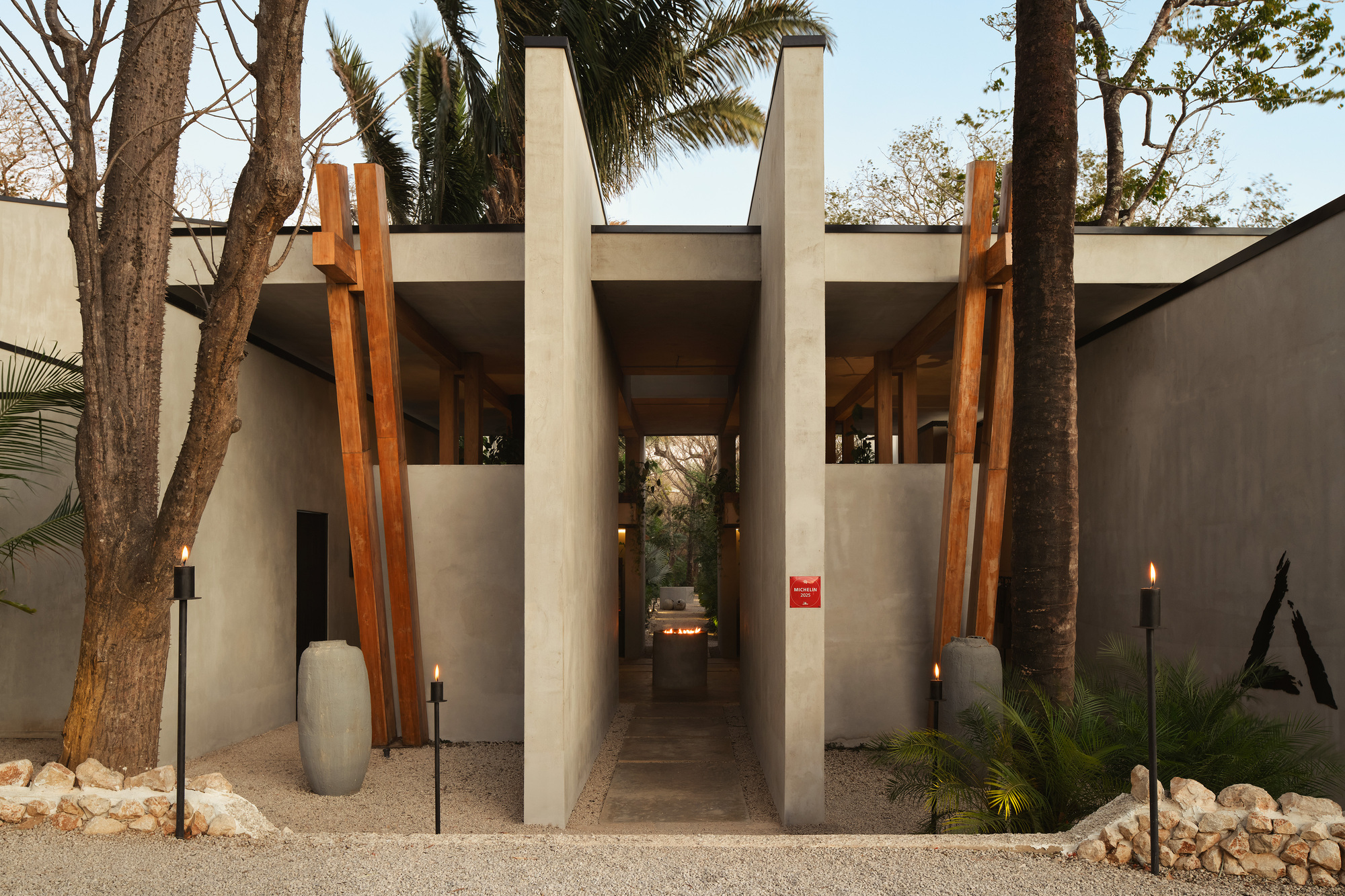 Gravel entry path between concrete walls and leaning timber posts, with lit torches and stacked stone edging in daylight