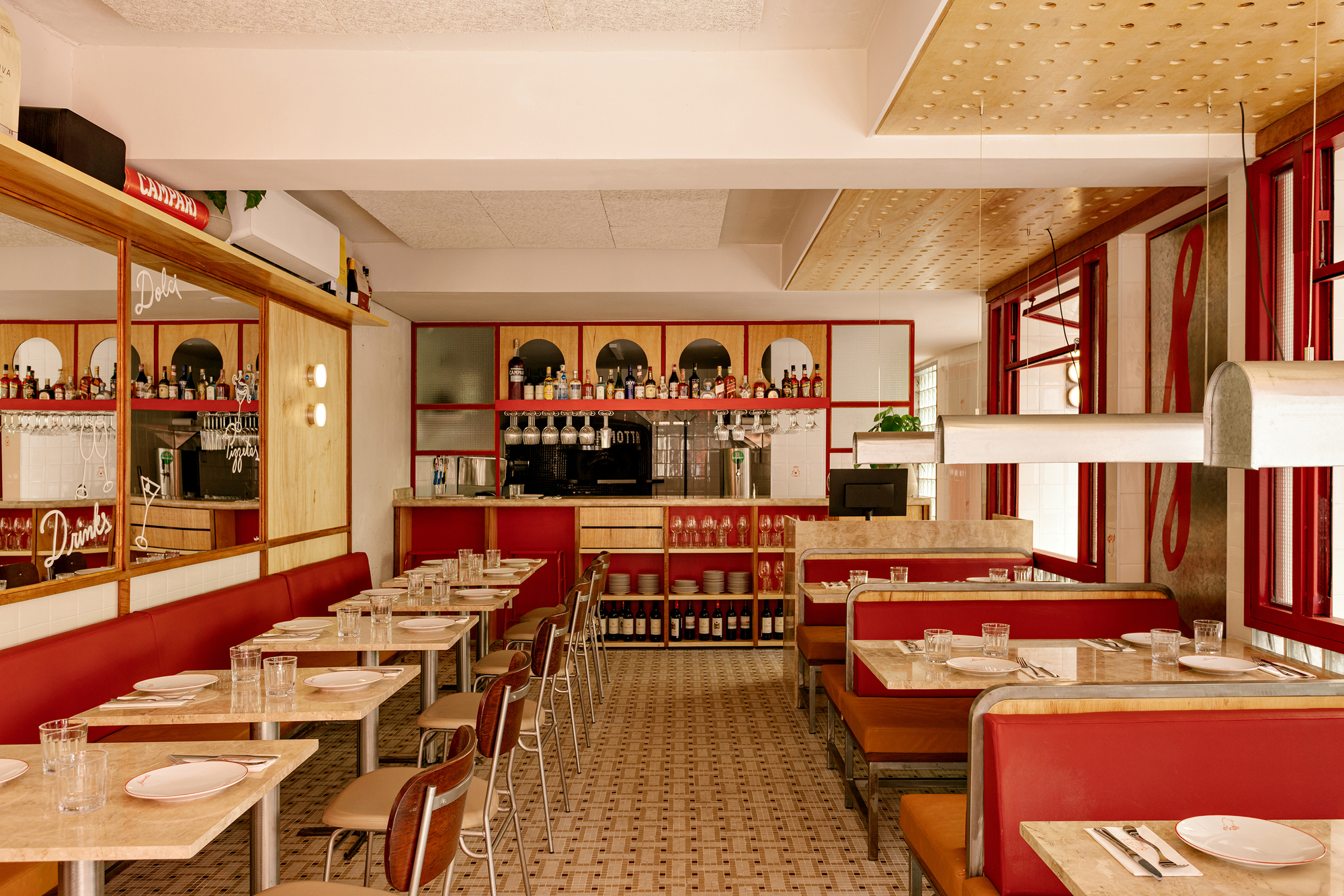 Dining room with red banquettes, stone-topped tables and a back bar of arched mirrors under perforated timber ceiling panels