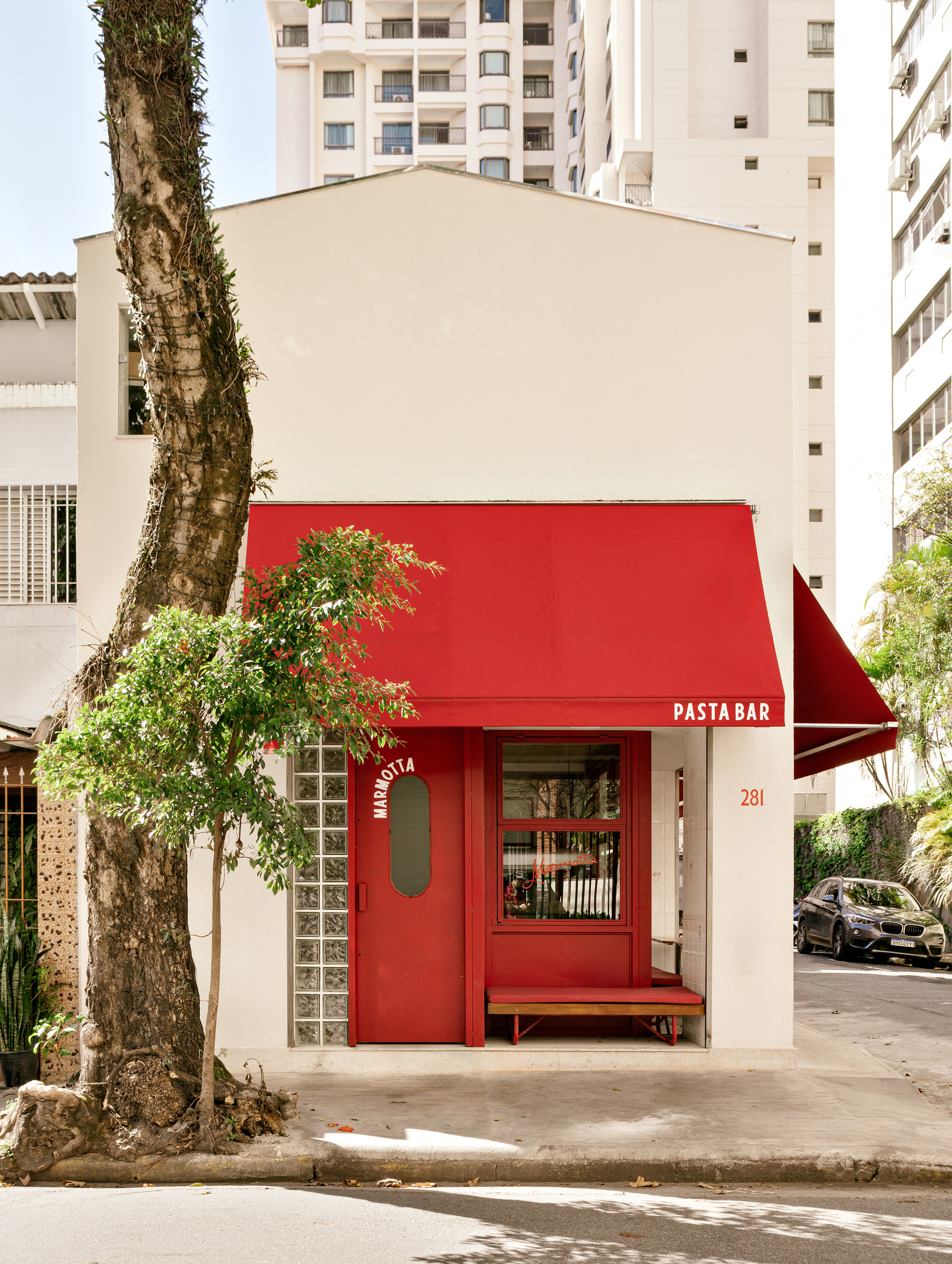 Narrow white gabled frontage with a red canvas awning, glass block panel and red door beside a street tree