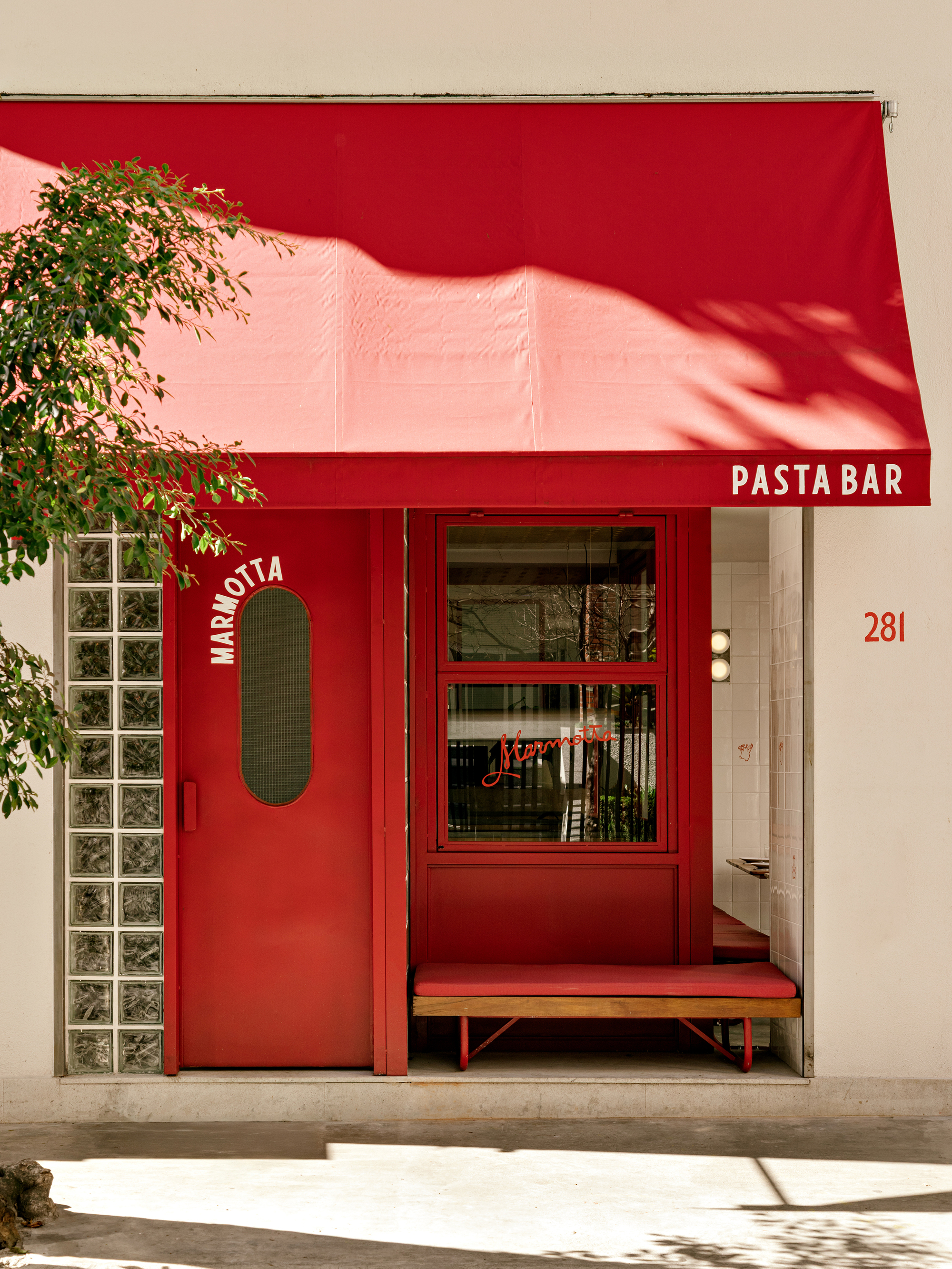 Close view of the red awning, oval-windowed door and glass block strip, with leaf shadows falling across the canvas