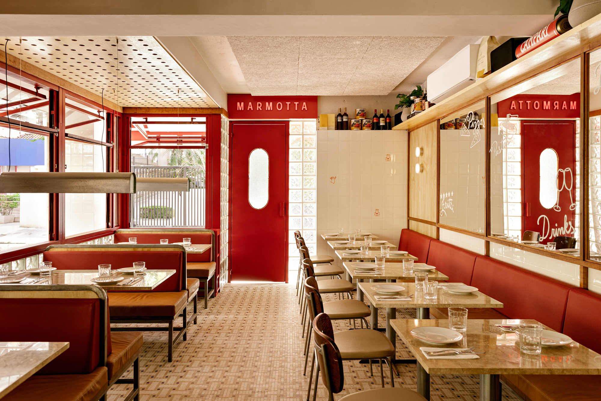 Long dining room looking toward a red door, with banquettes, stone tables and patterned floor tile in daylight