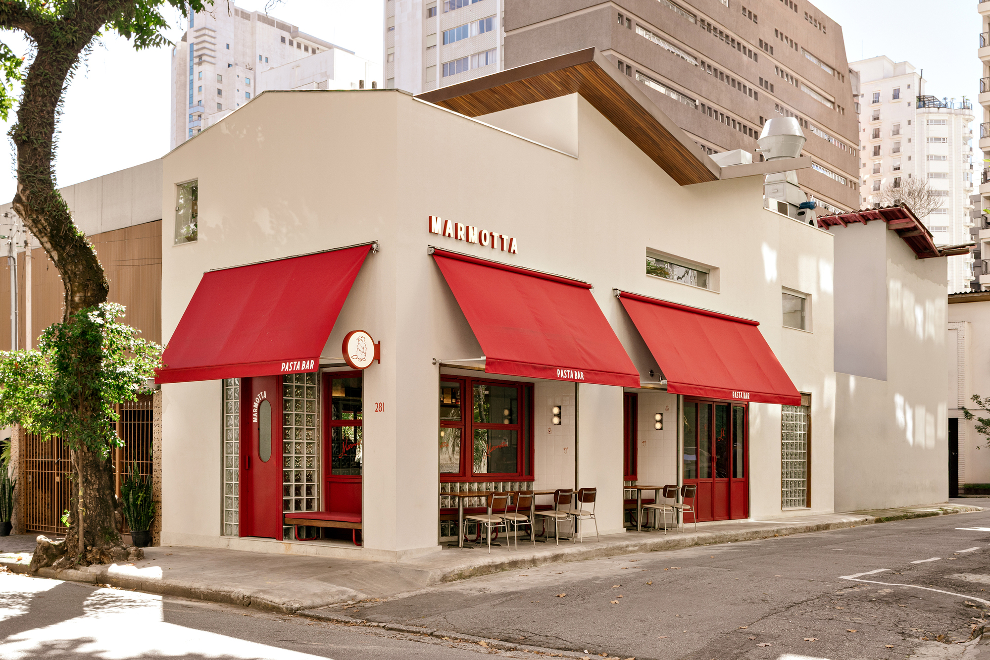 Corner view of the white facade with three red awnings and sidewalk tables, set against apartment towers in afternoon sun