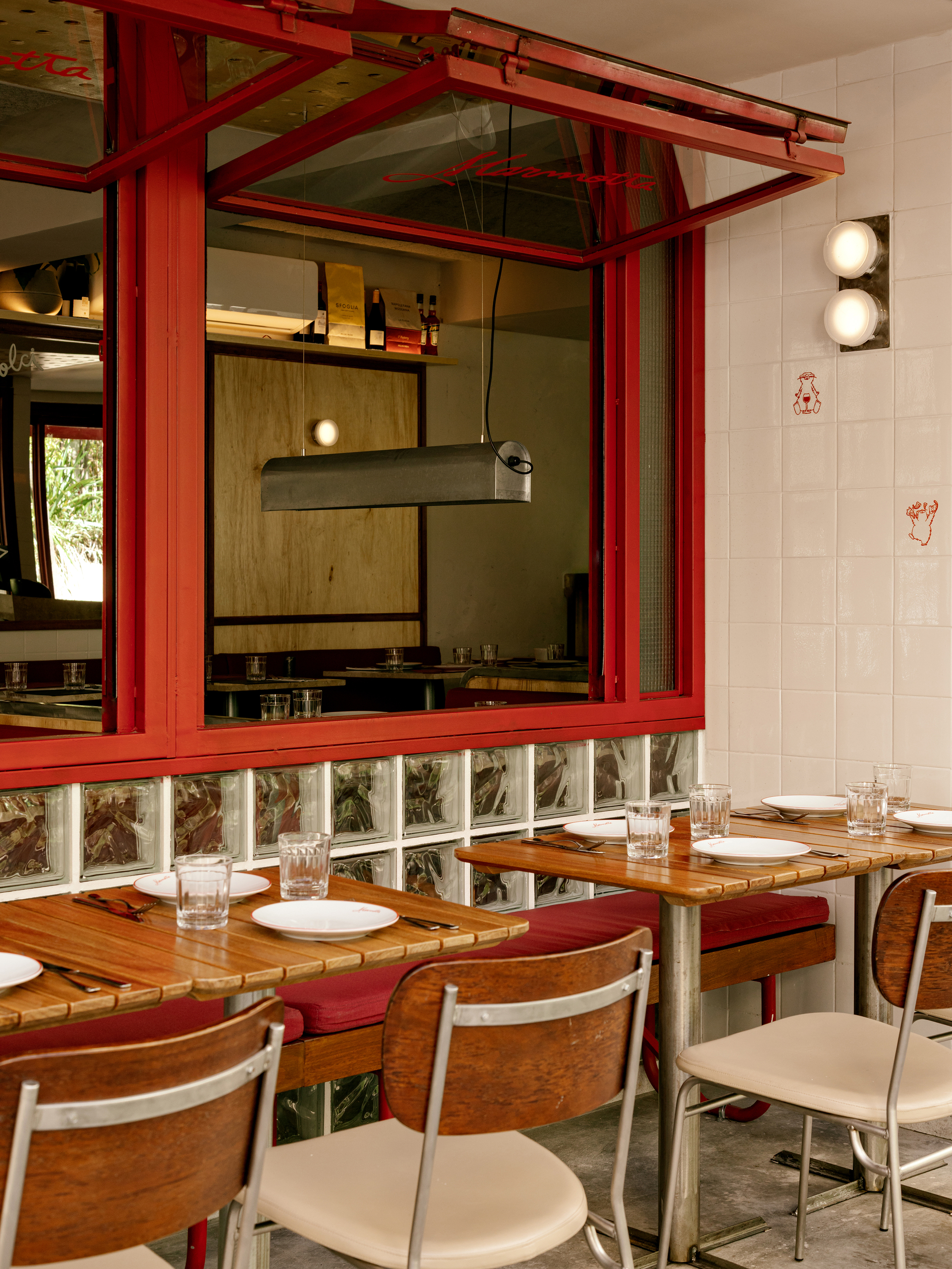 Red-framed pivot window above a glass block base, with wood-top tables and chrome chairs set for lunch in daylight