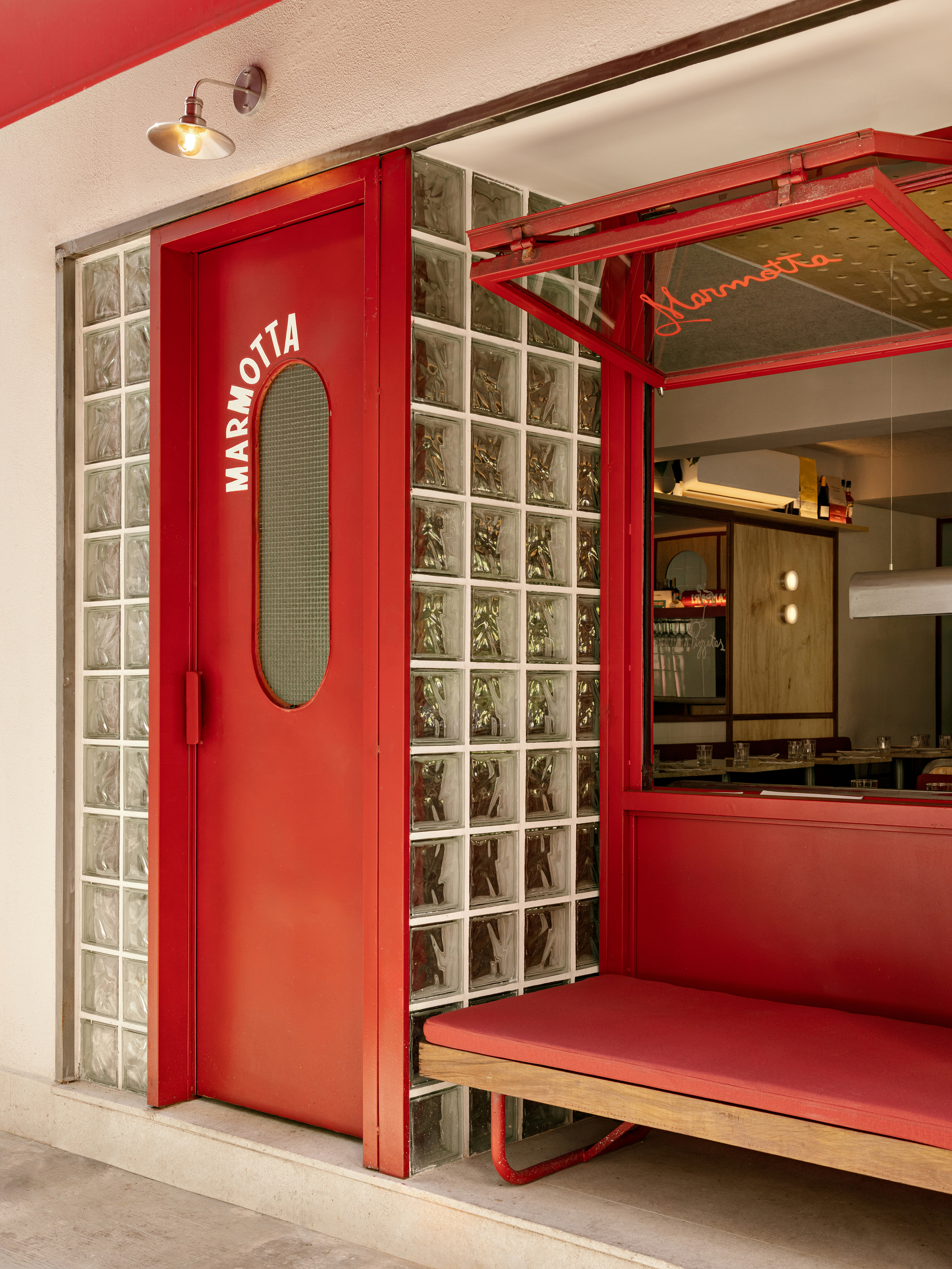 Red entry door with oval mesh glass beside a glass block wall and a cushioned red bench under a lamp