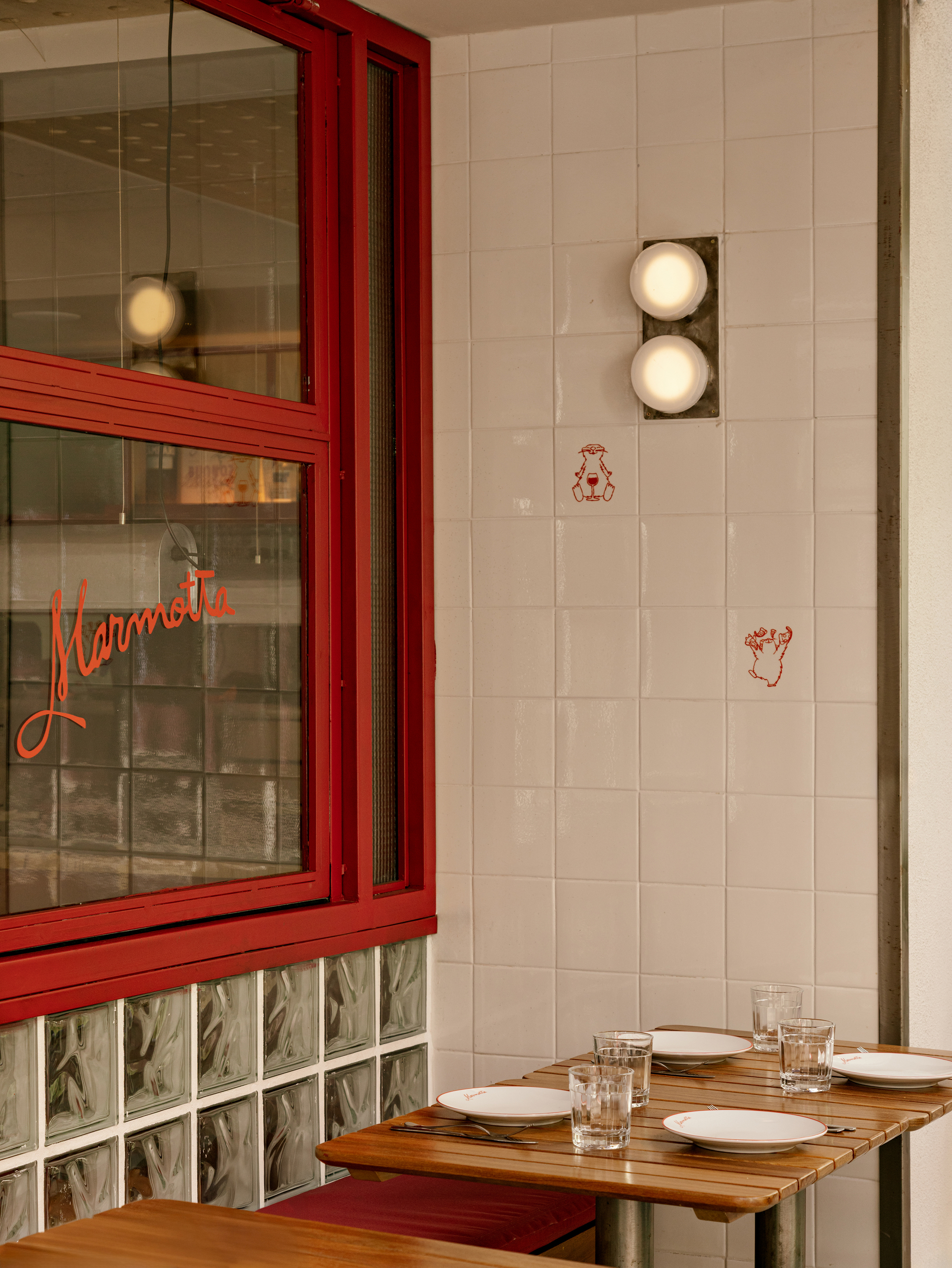 Corner of white tile wall with small red line drawings, twin globe sconces and a set wooden table in soft light