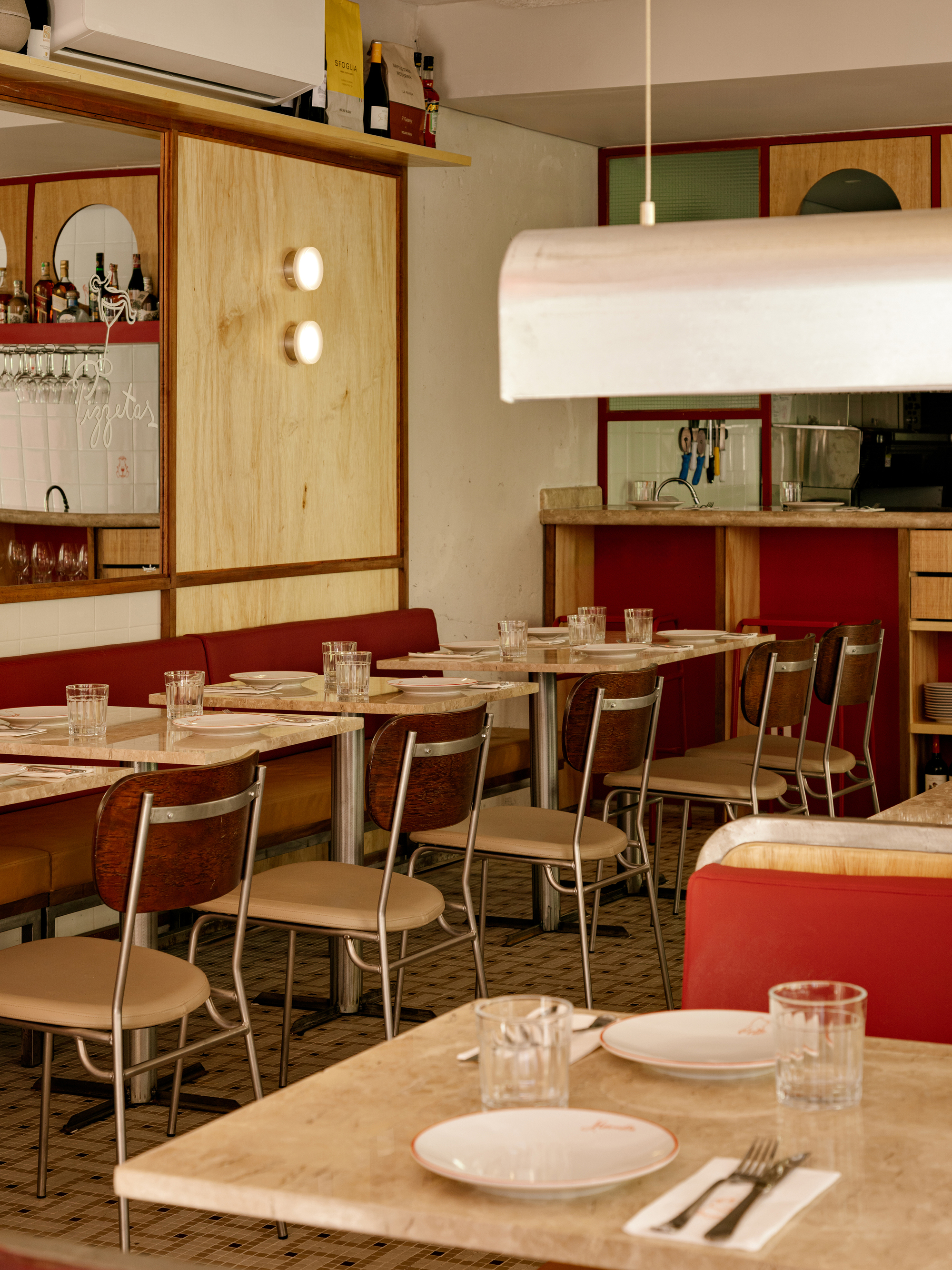 Dining room with red banquettes, plywood panelled bar back and steel-framed chairs over a small checked tile floor