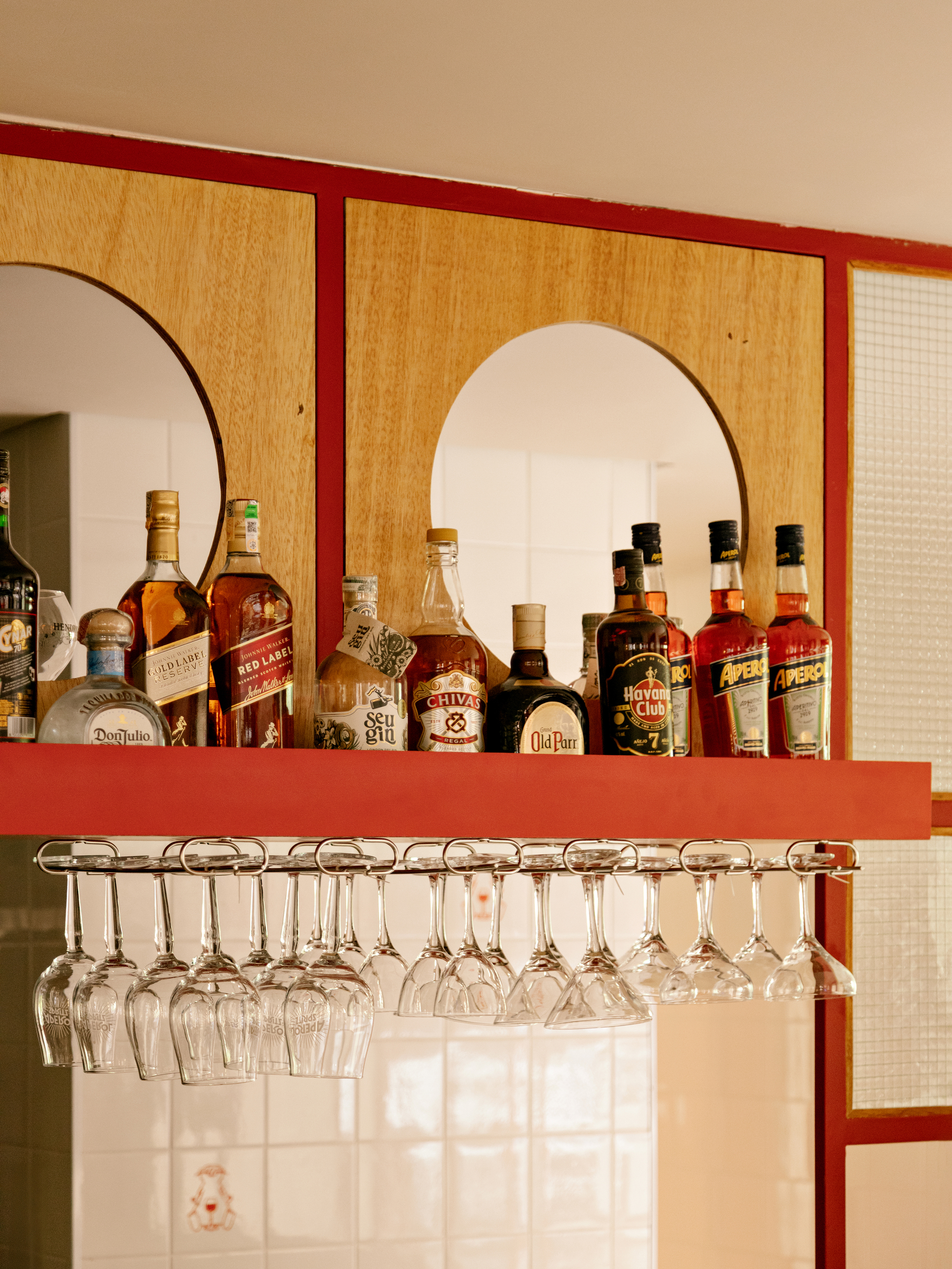 Red shelf with liquor bottles below arched mirror cutouts in plywood, hanging wine glasses above white tile and reeded glass in warm light