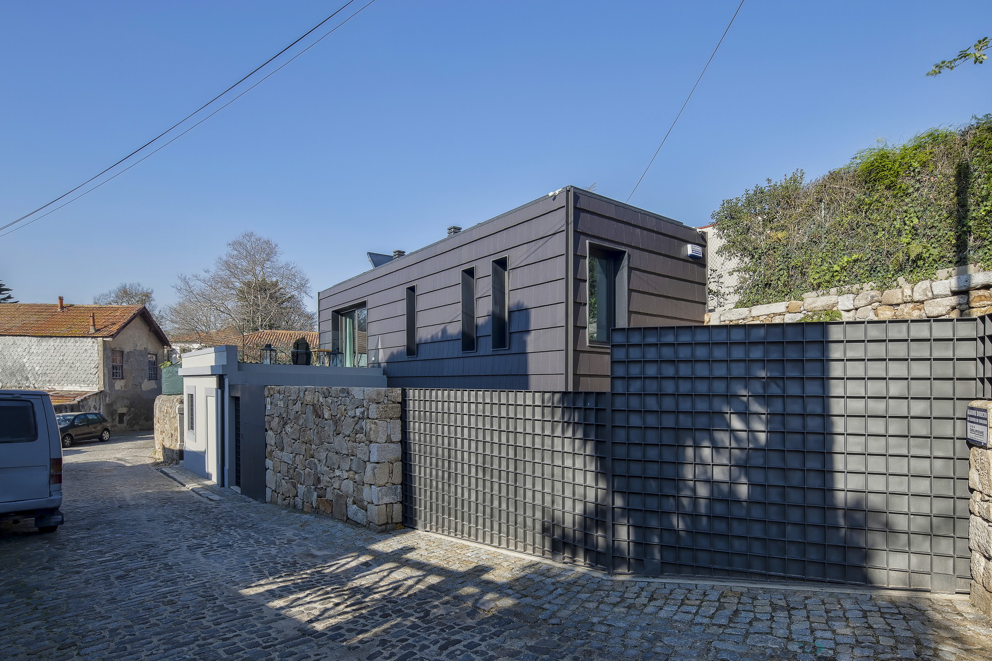Street corner with dark grid-patterned gate, rough stone wall and slate-clad upper volume on cobbles in clear afternoon sun
