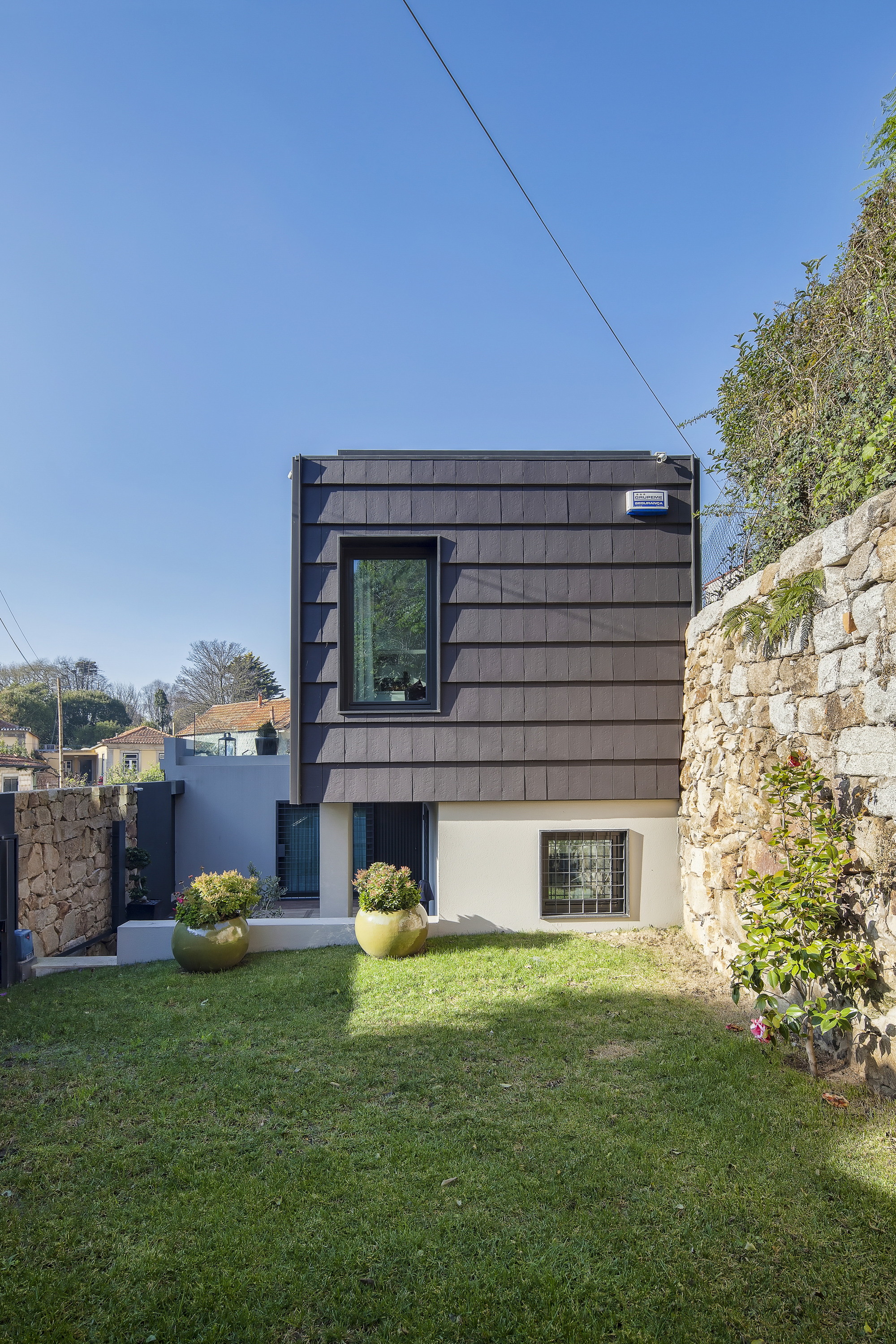 Rear garden facade with slate-clad upper box above a white base, flanked by a stone wall and two potted shrubs