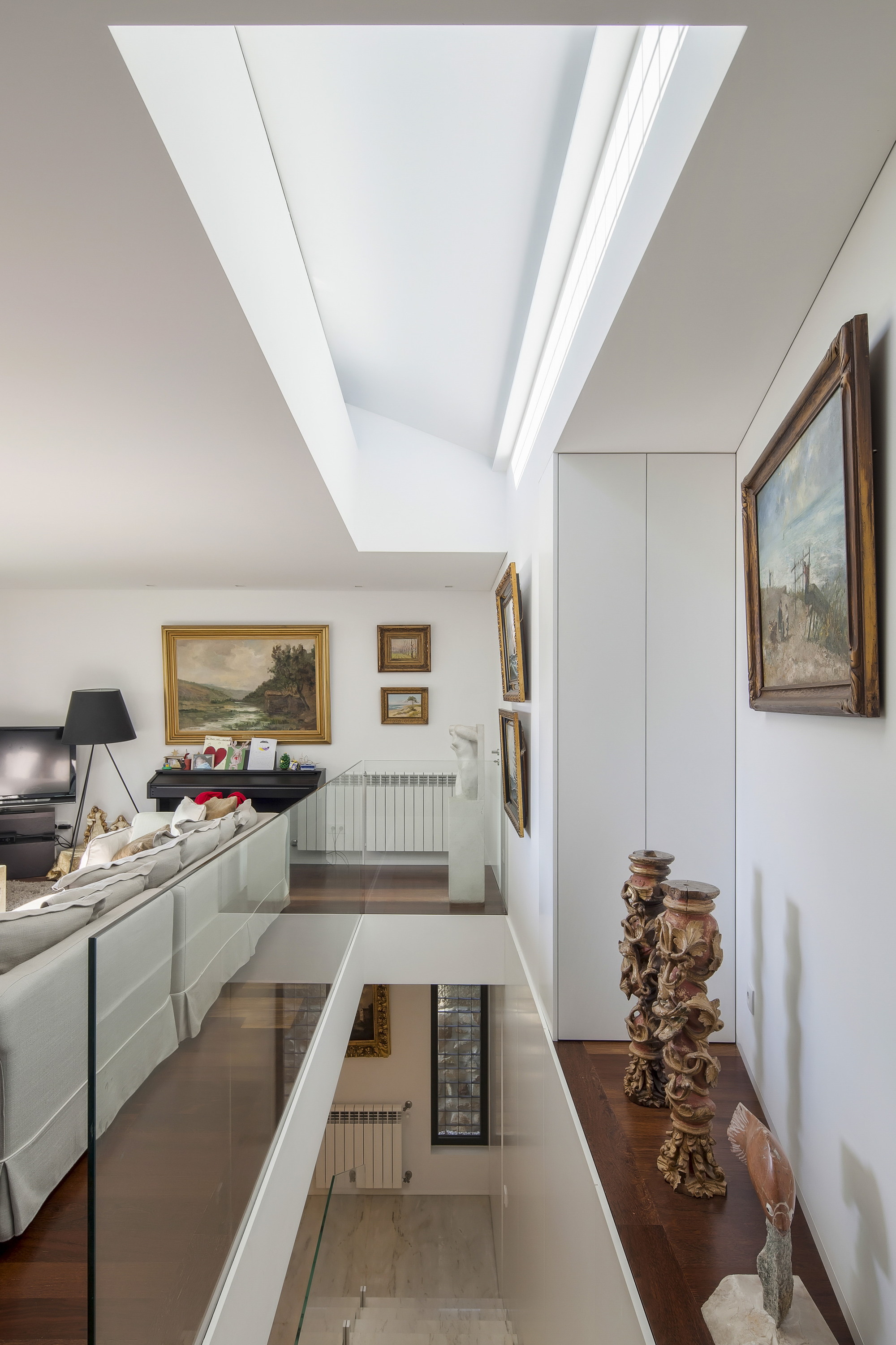 Upper floor landing with glass balustrade beside a skylight well, framed paintings and carved wooden columns on a dark timber ledge
