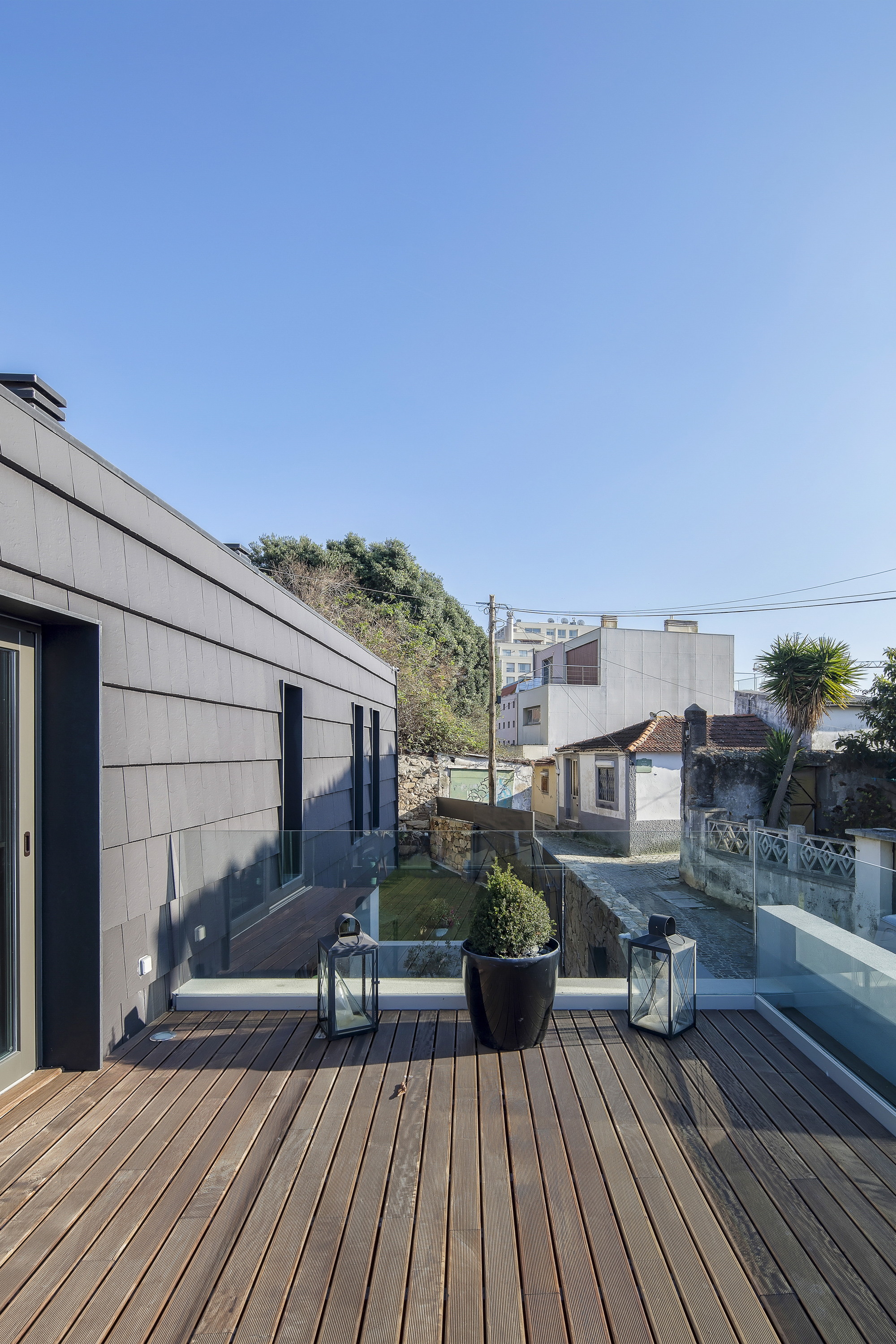 Timber roof terrace with a potted shrub and two lanterns beside slate-clad walls, overlooking neighbouring rooftops under a clear sky