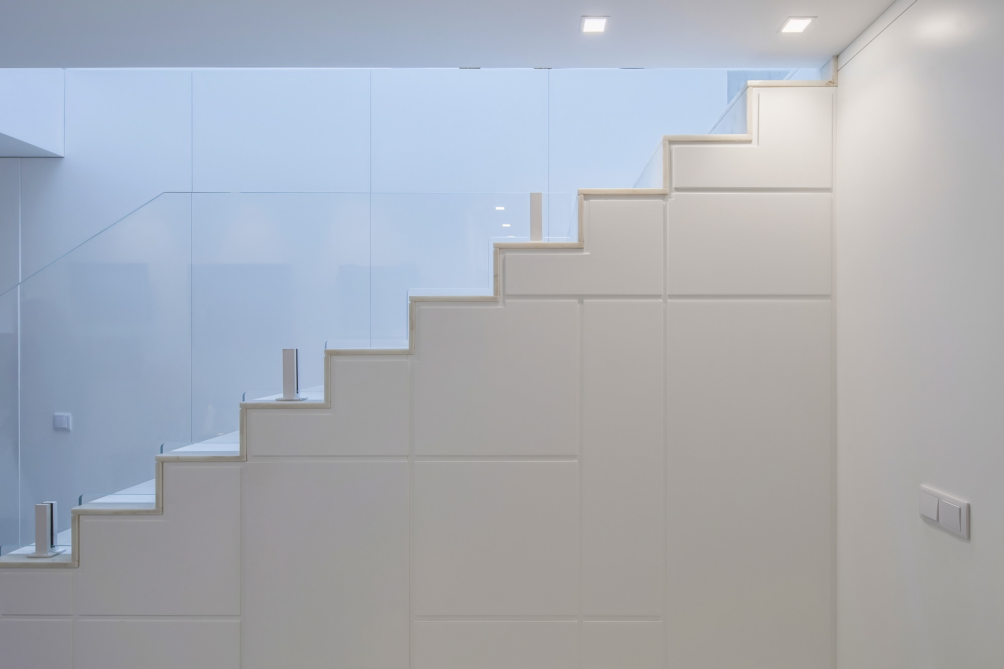 Side view of a white stair with panelled cladding and glass balustrade, lit by recessed ceiling spotlights and cool daylight