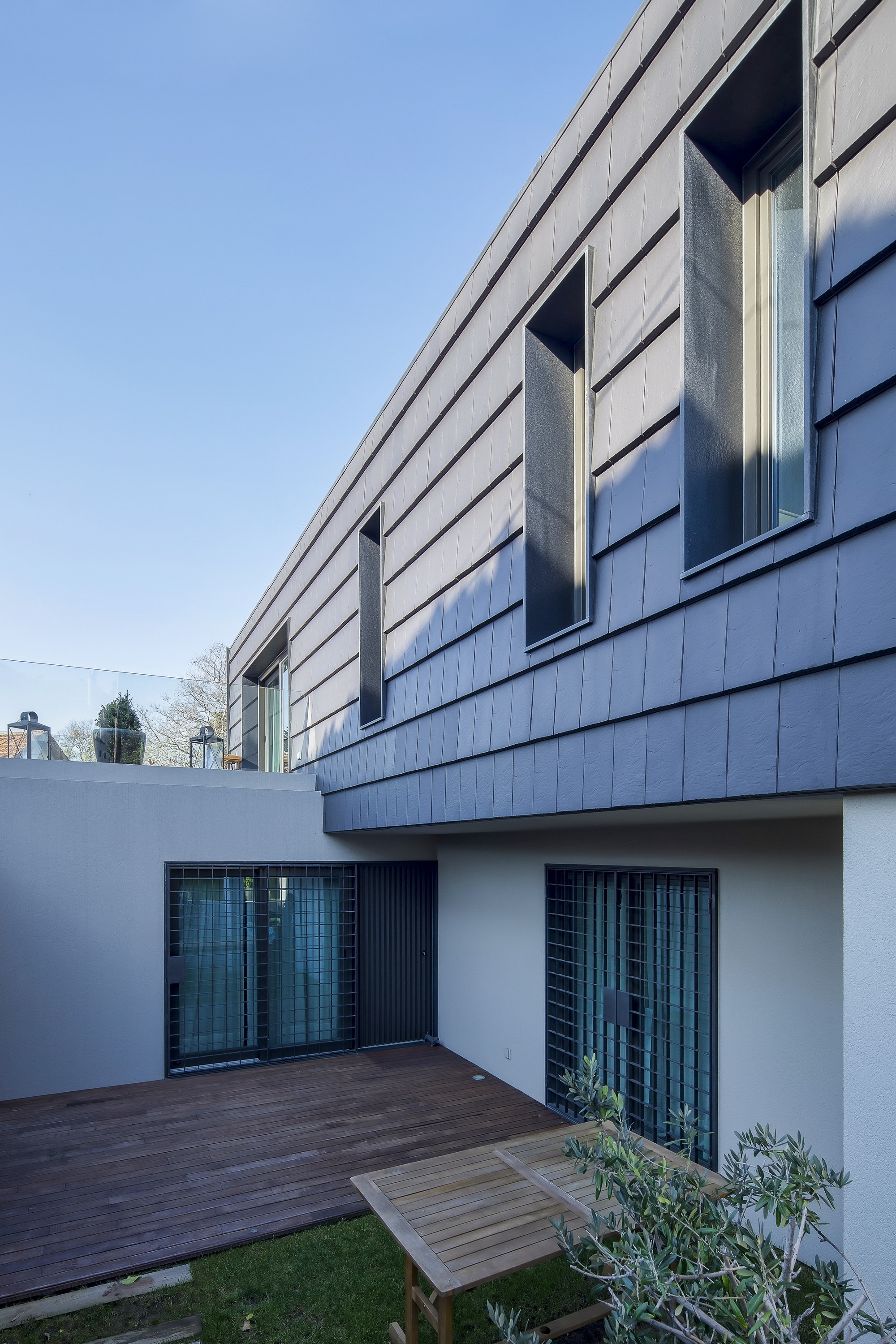 Courtyard with a timber deck and wooden table beneath the slate-clad upper floor, tall recessed windows catching low afternoon sun