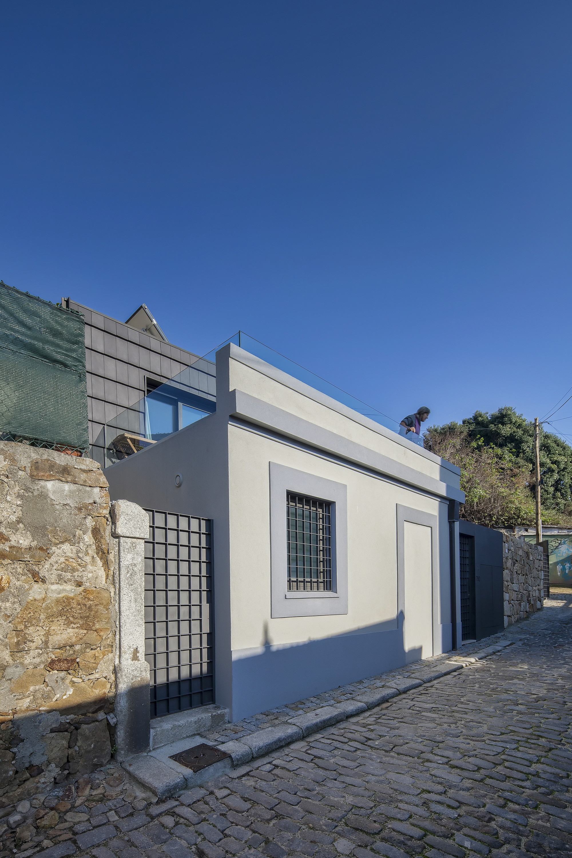 Pale grey rendered street facade with a barred window, beside a rough stone wall and cobbled lane under clear blue sky