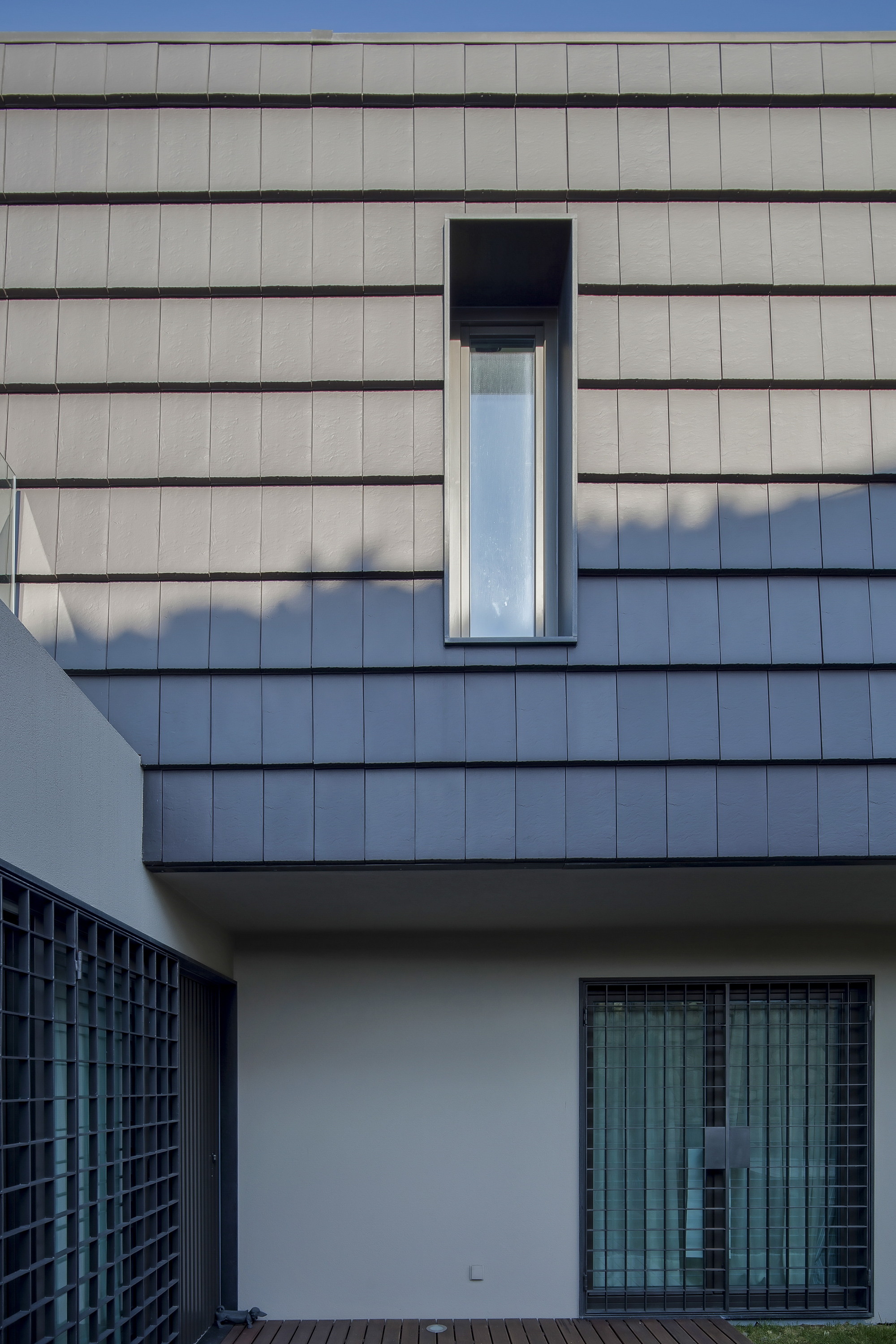 Overlapping grey cladding shingles on the upper volume with a tall narrow window, crossed by tree shadow in afternoon light