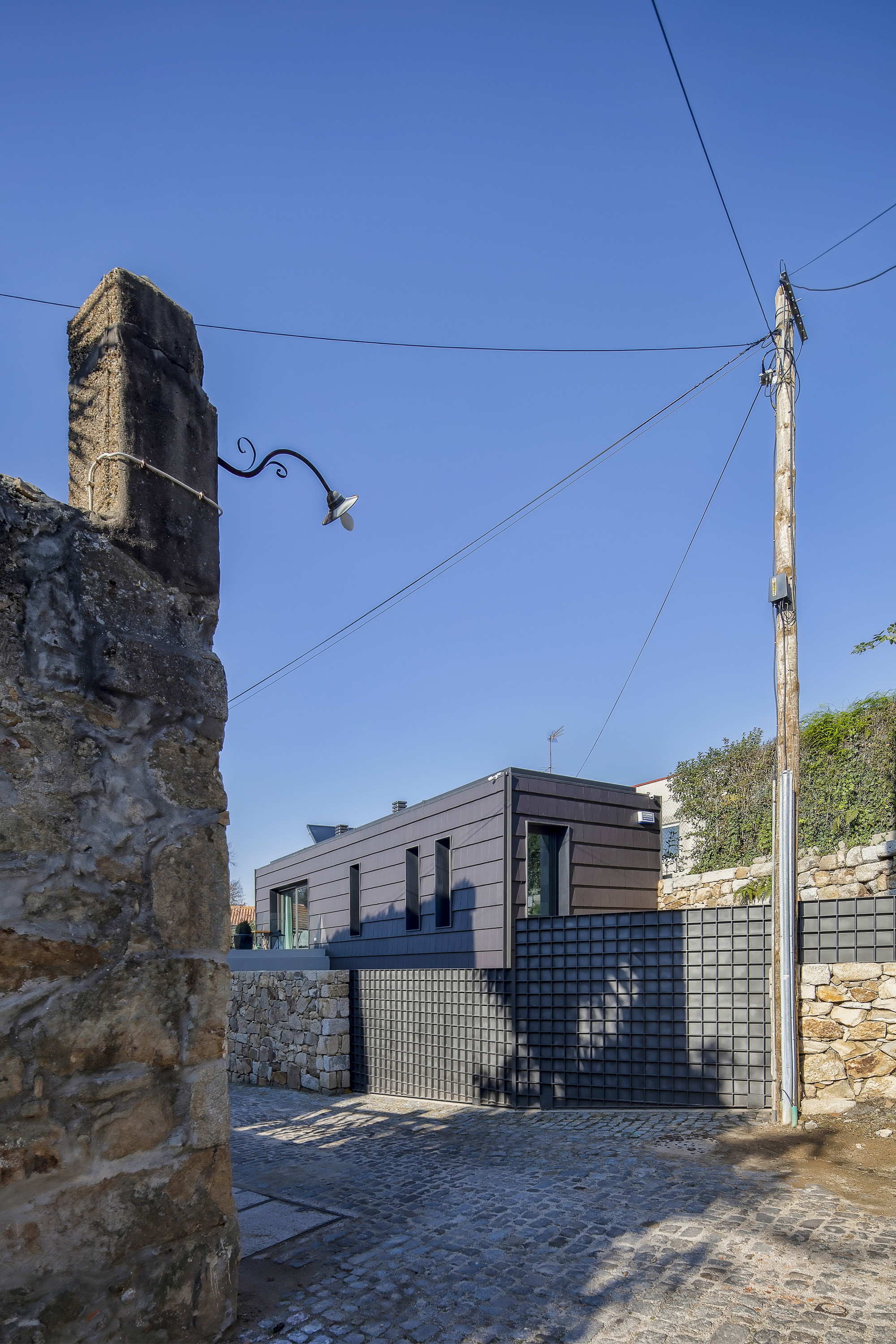 Dark cladded house behind a gridded metal gate and stone walls, seen past a weathered gatepost and utility pole