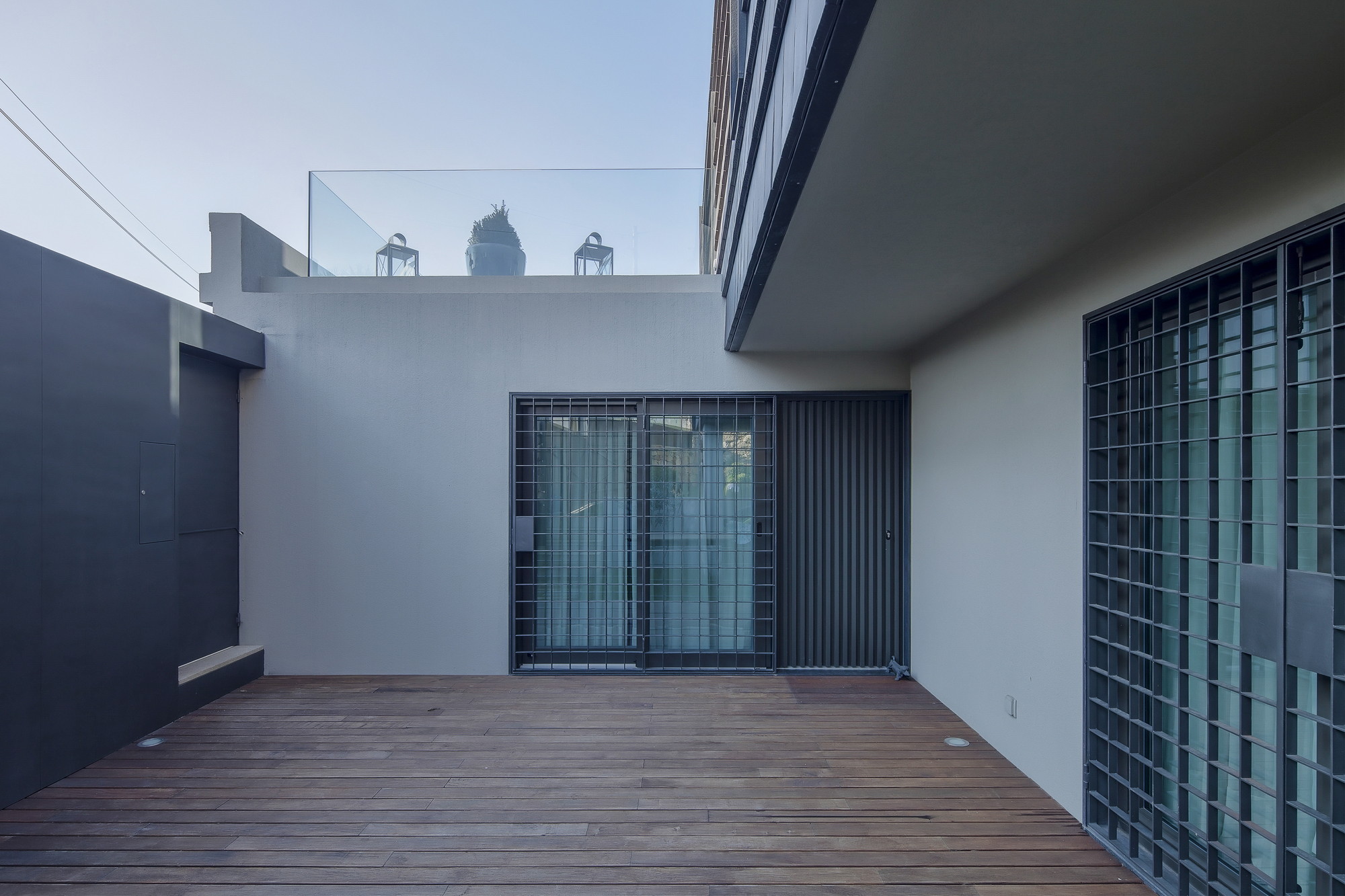 Timber deck courtyard enclosed by grey walls and gridded steel doors, with a glass-railed roof terrace above at dusk