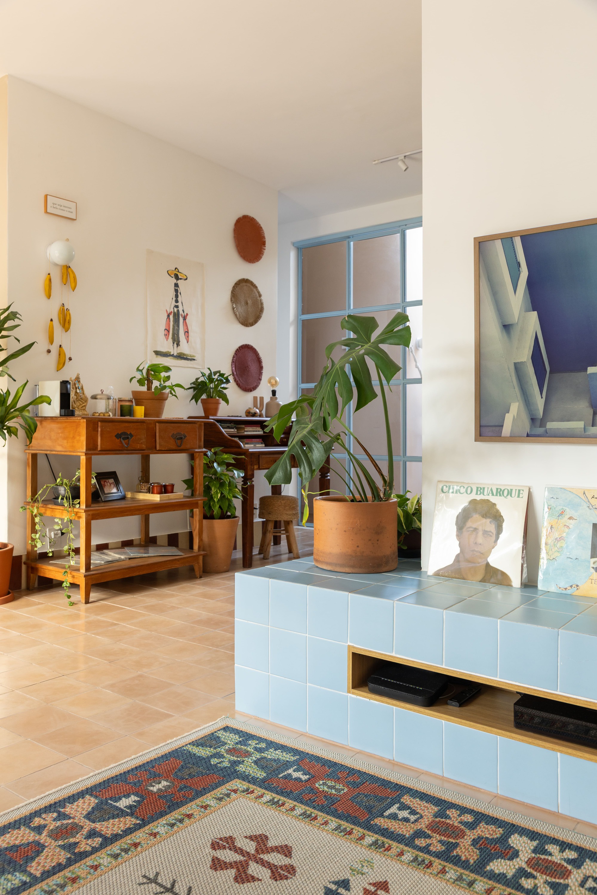Blue tiled bench with a monstera pot and vinyl records, beside a wooden console and woven wall plates in warm light