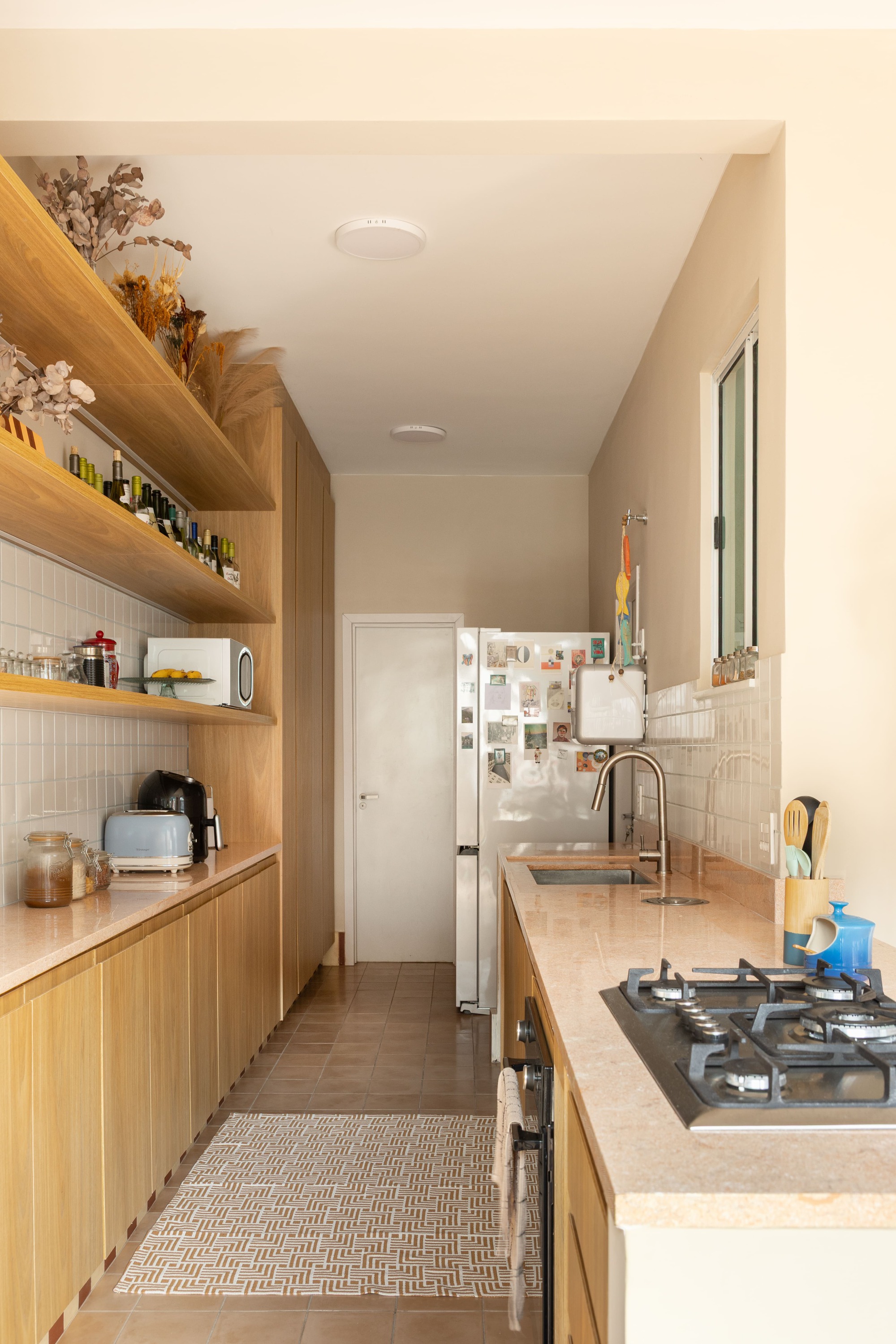Galley kitchen with oak cabinetry, open shelves, pink stone countertop and a patterned rug on the terracotta floor