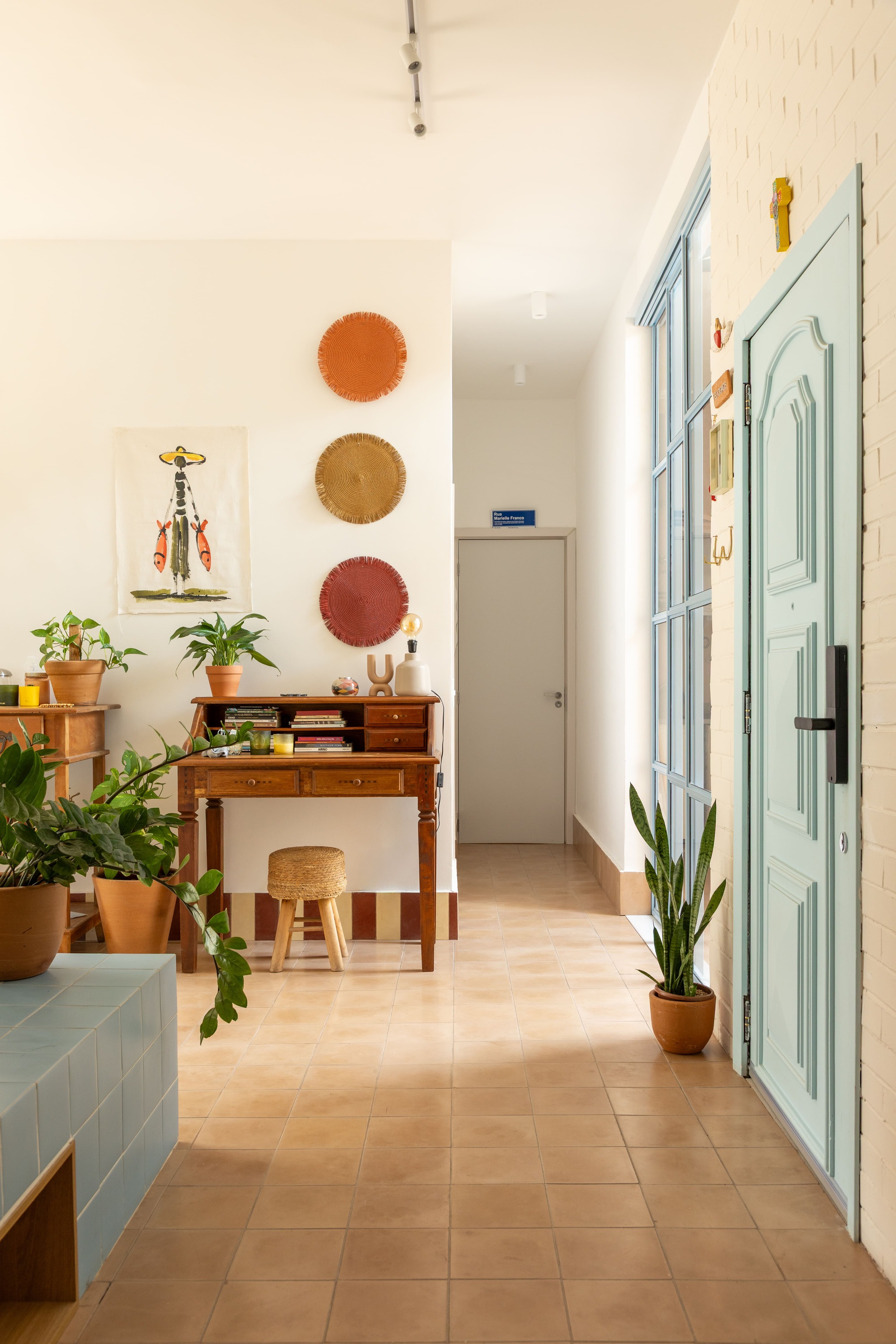 Hallway view toward a wooden writing desk with three woven wall discs, a stool and a pale blue door in afternoon sun