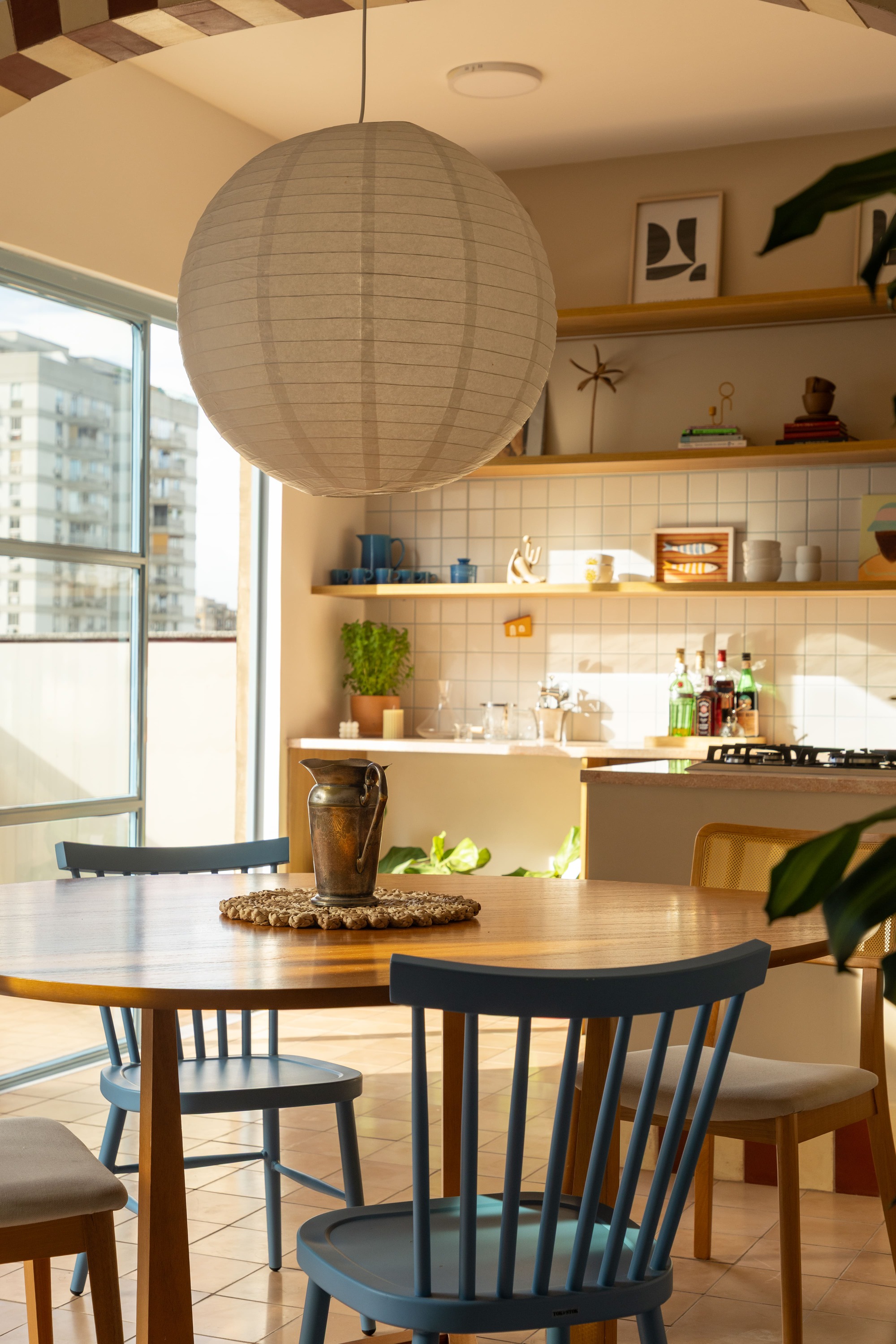 Paper lantern pendant above the dining table, with open shelves and a tiled backsplash lit by low afternoon sun