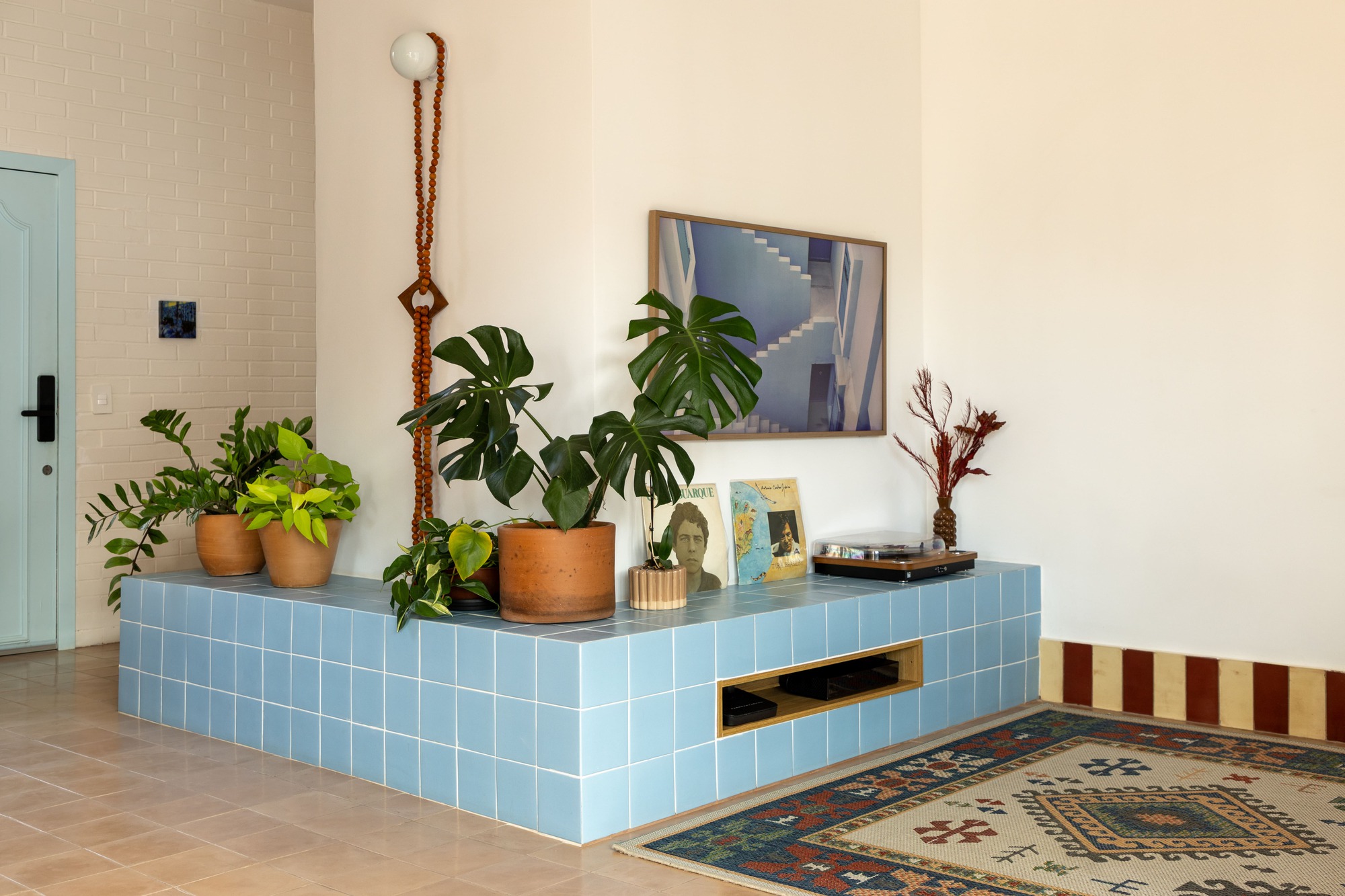 Blue tiled platform holding potted monstera plants and a record player, beside a pale blue door and a patterned rug