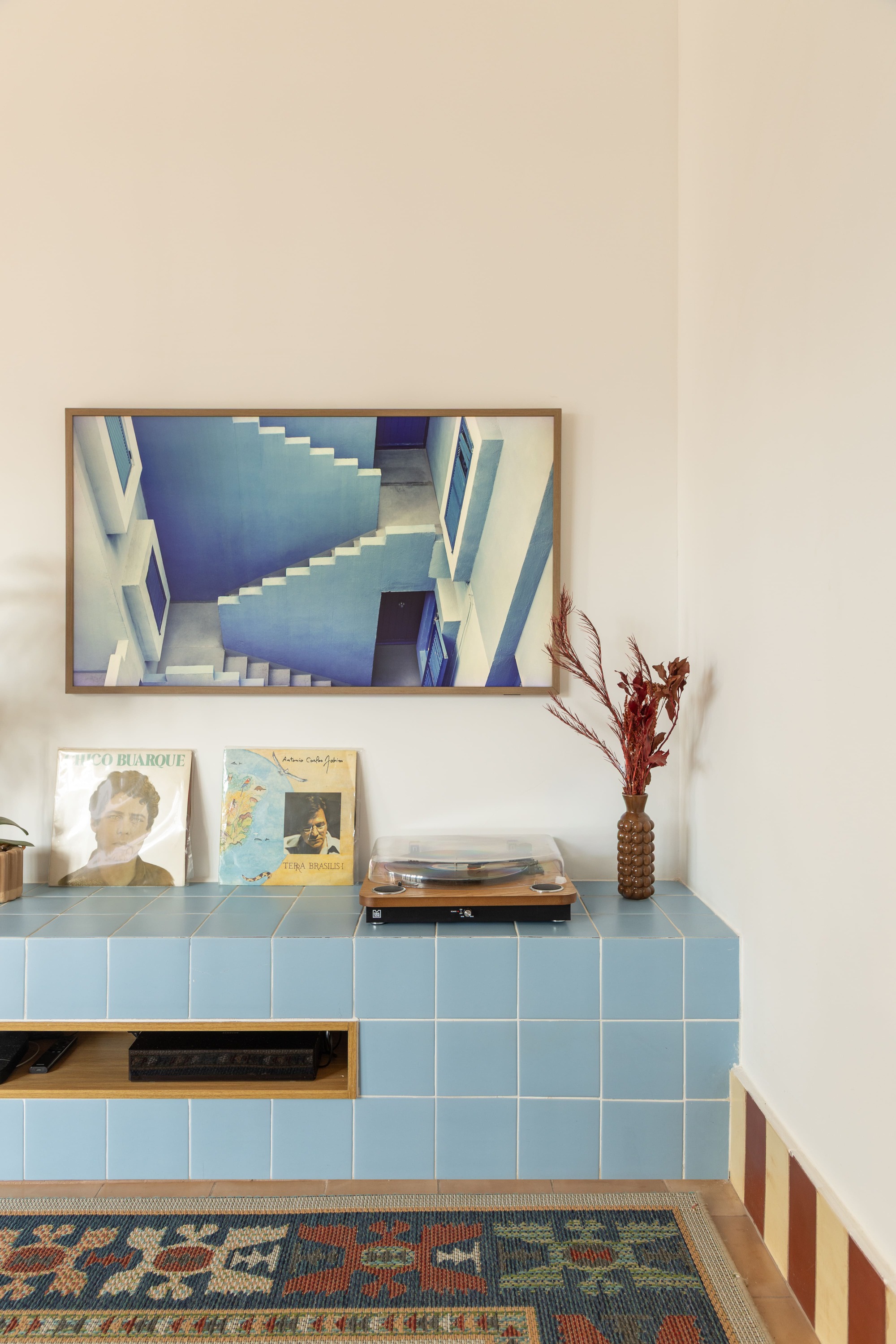 Pale blue tiled ledge holding a turntable and vinyl records beneath a framed print of blue stairs, with dried flowers in a vase