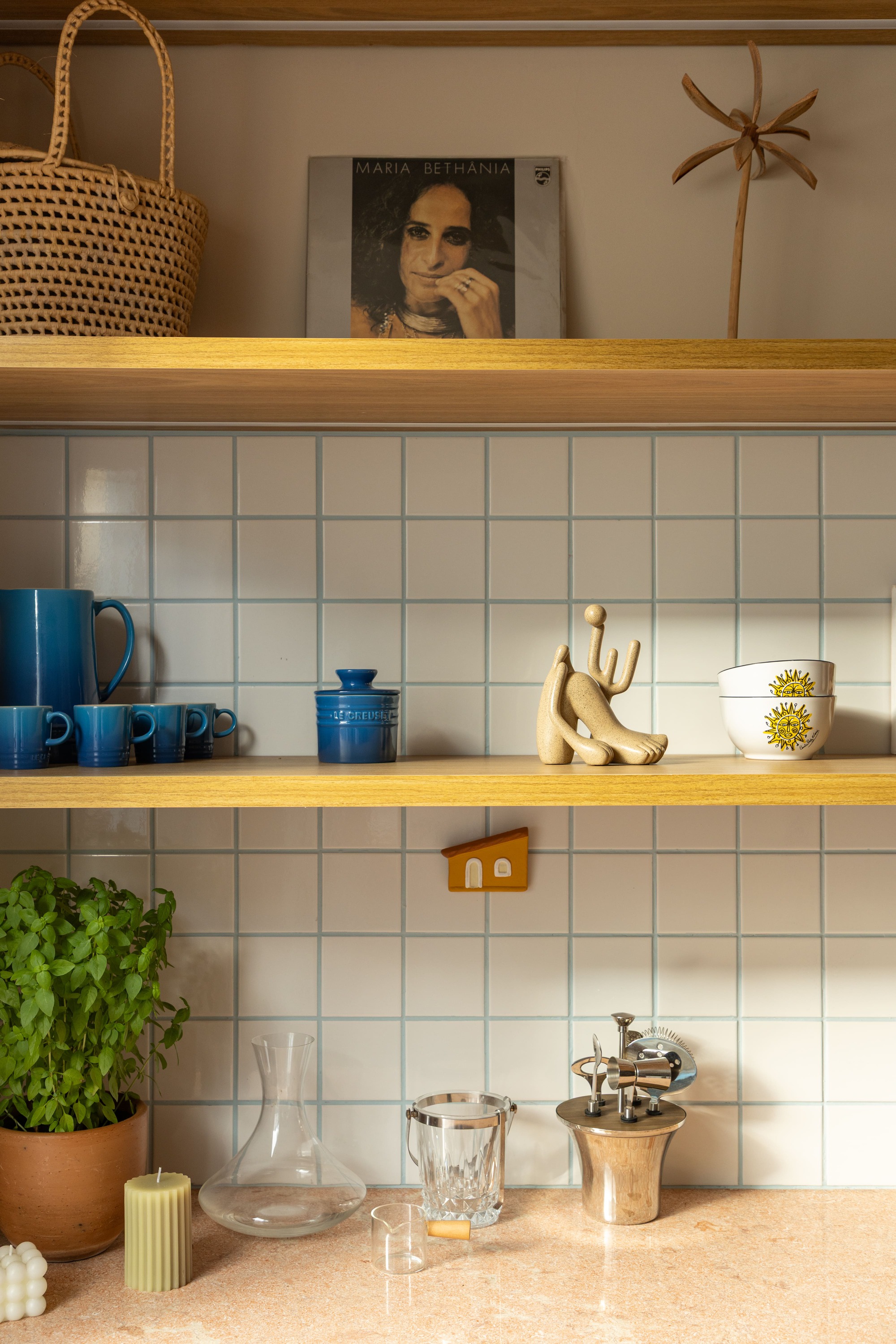Oak shelves over pale tile backsplash holding blue mugs, a sculpture, a basil plant and a glass carafe on a pink stone counter
