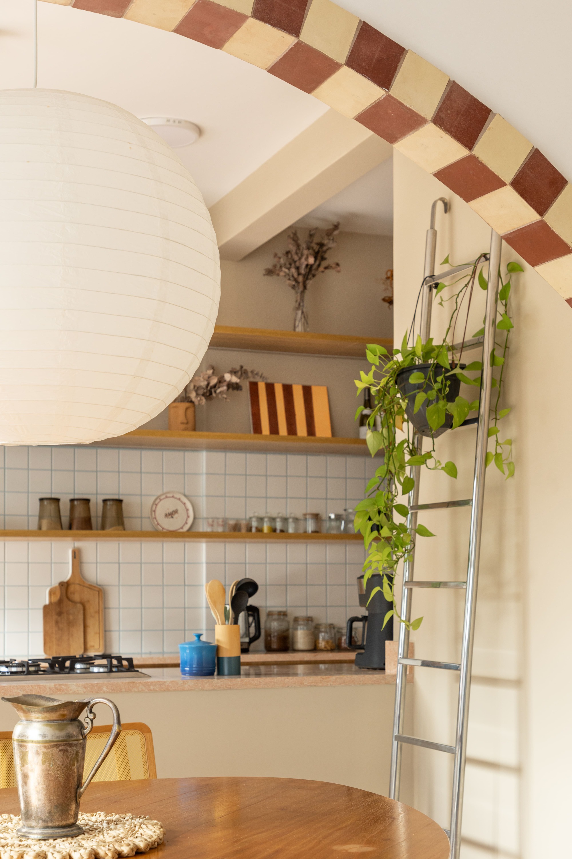 Kitchen seen through an arch trimmed in checkered terracotta and cream tile, with a paper lantern and a metal ladder hung with trailing plants