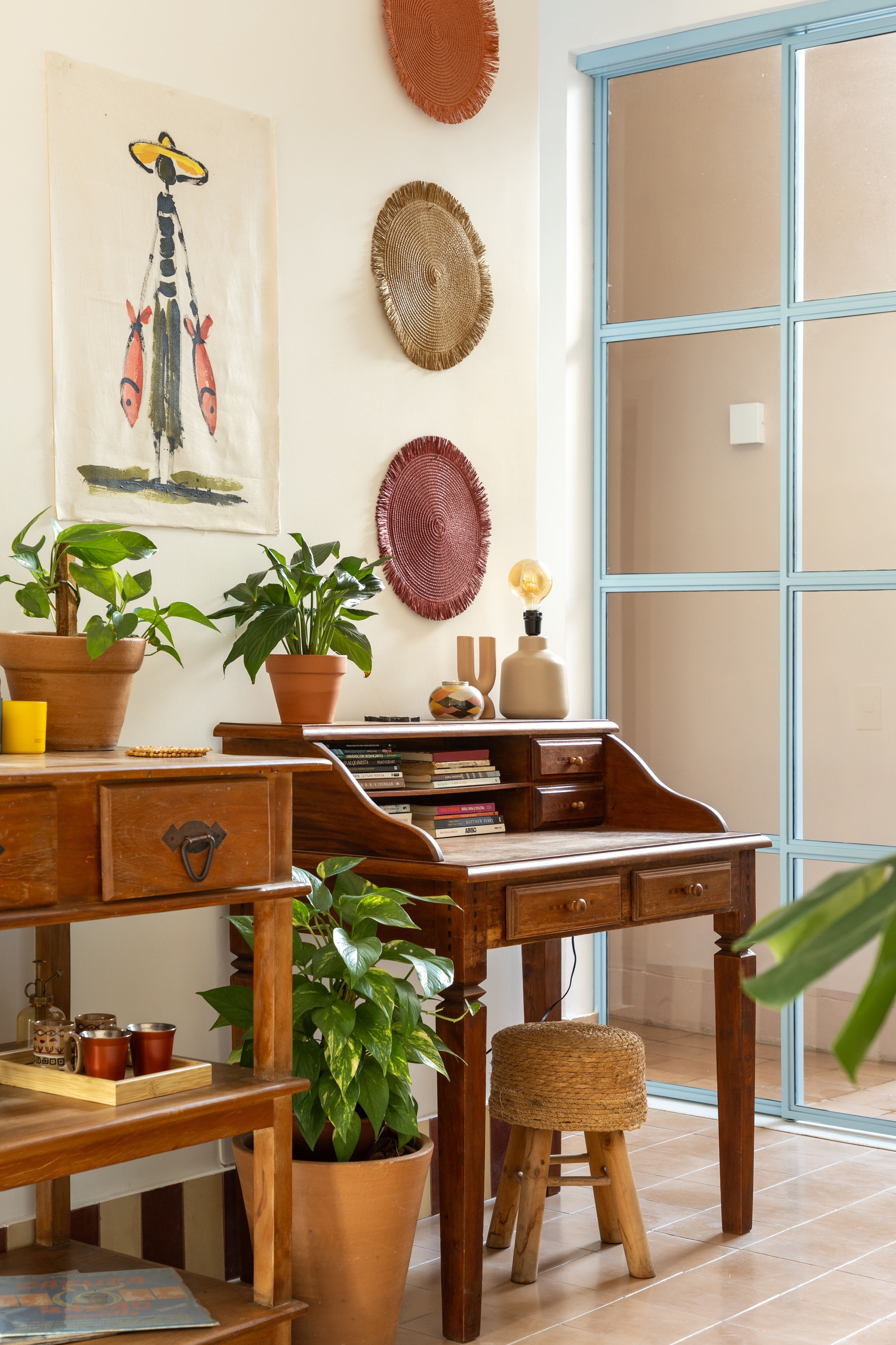 Wooden writing desk with potted plants and woven wall plates beside a pale blue steel-framed glass door in soft daylight