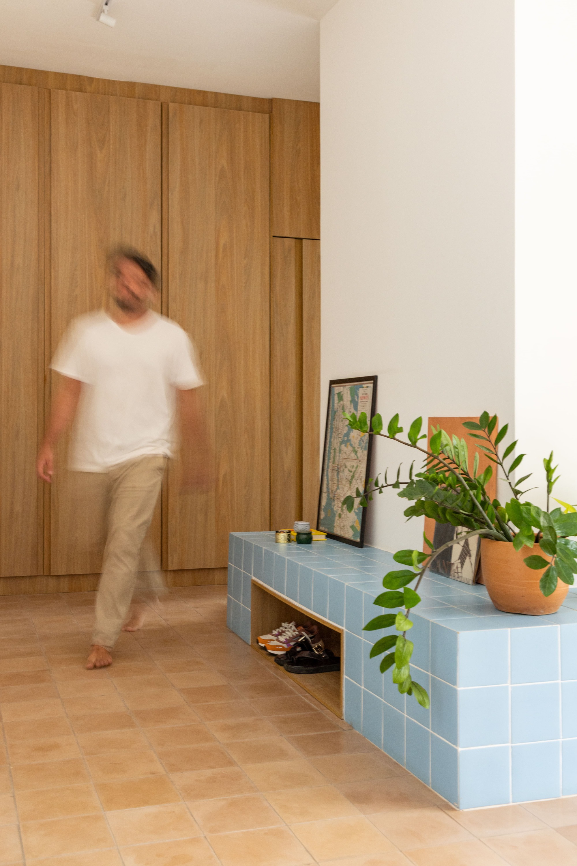 Barefoot man in motion blur crossing a terracotta tile floor past walnut cabinetry and a blue tiled bench with a potted plant