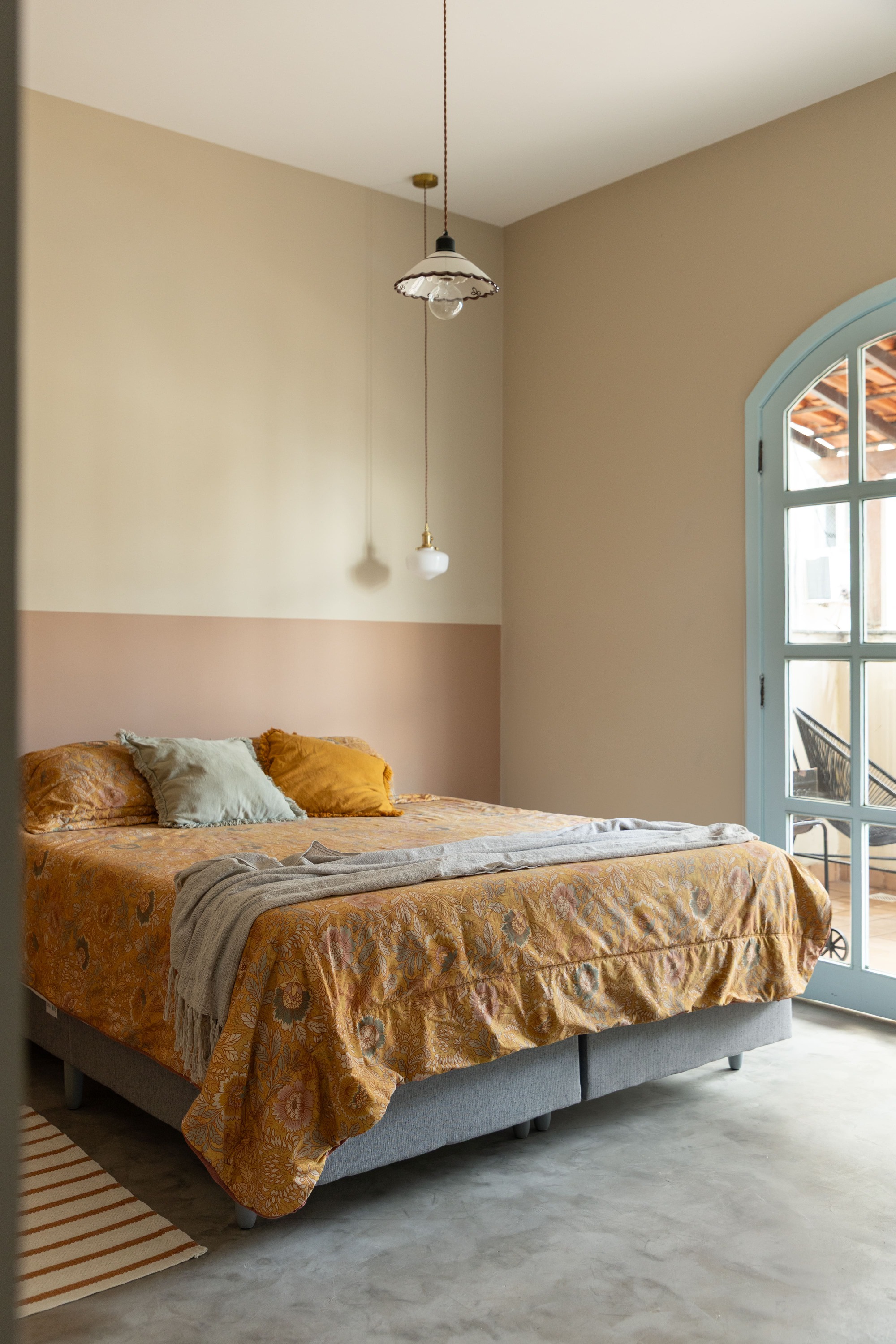Bedroom with a half-pink wall, ochre patterned bedspread, pendant lamps and an arched pale blue window door opening to a terrace