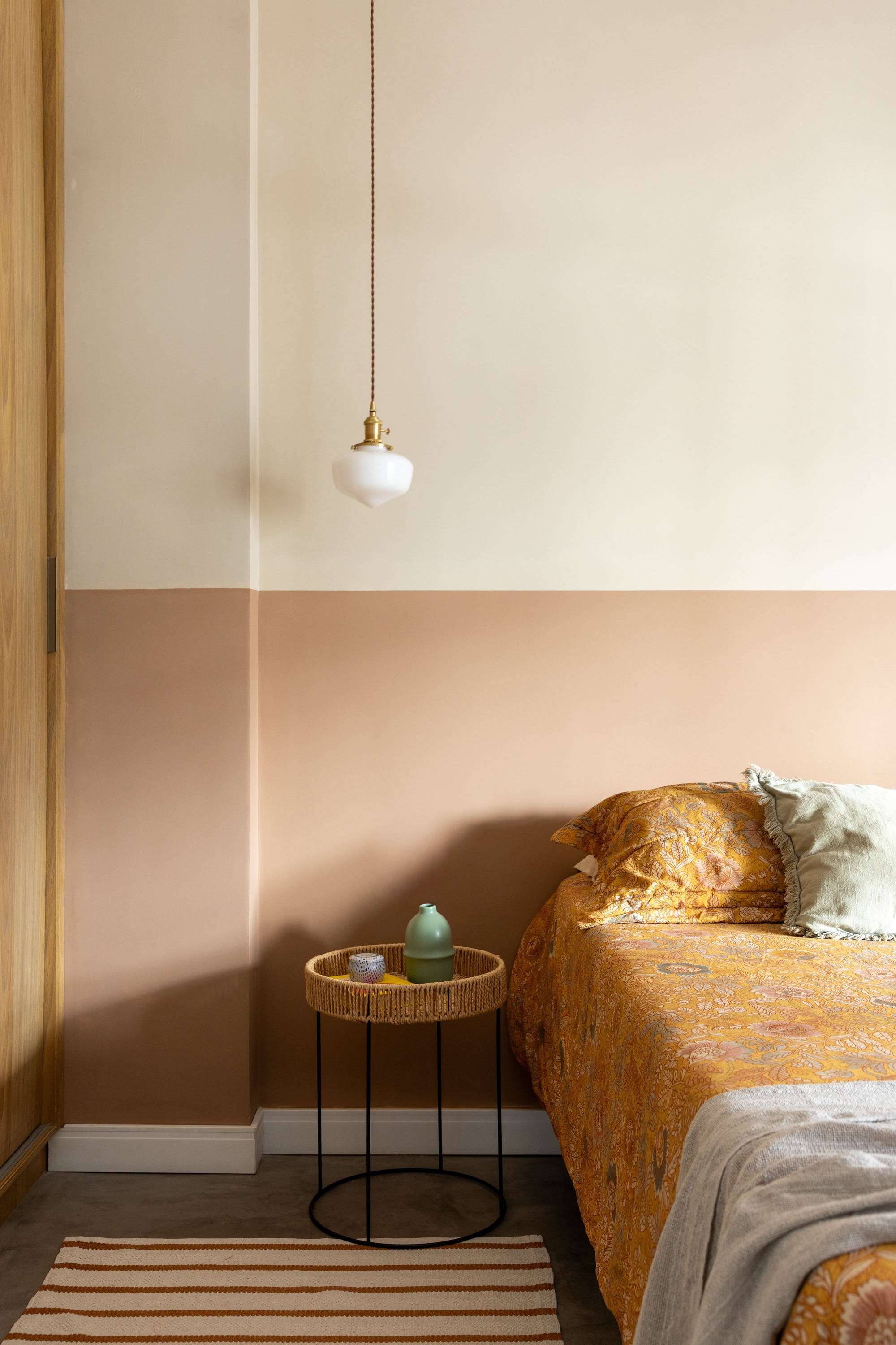 Bedroom corner with a two-tone cream and blush wall, woven side table, glass pendant lamp and ochre floral bedding in soft daylight