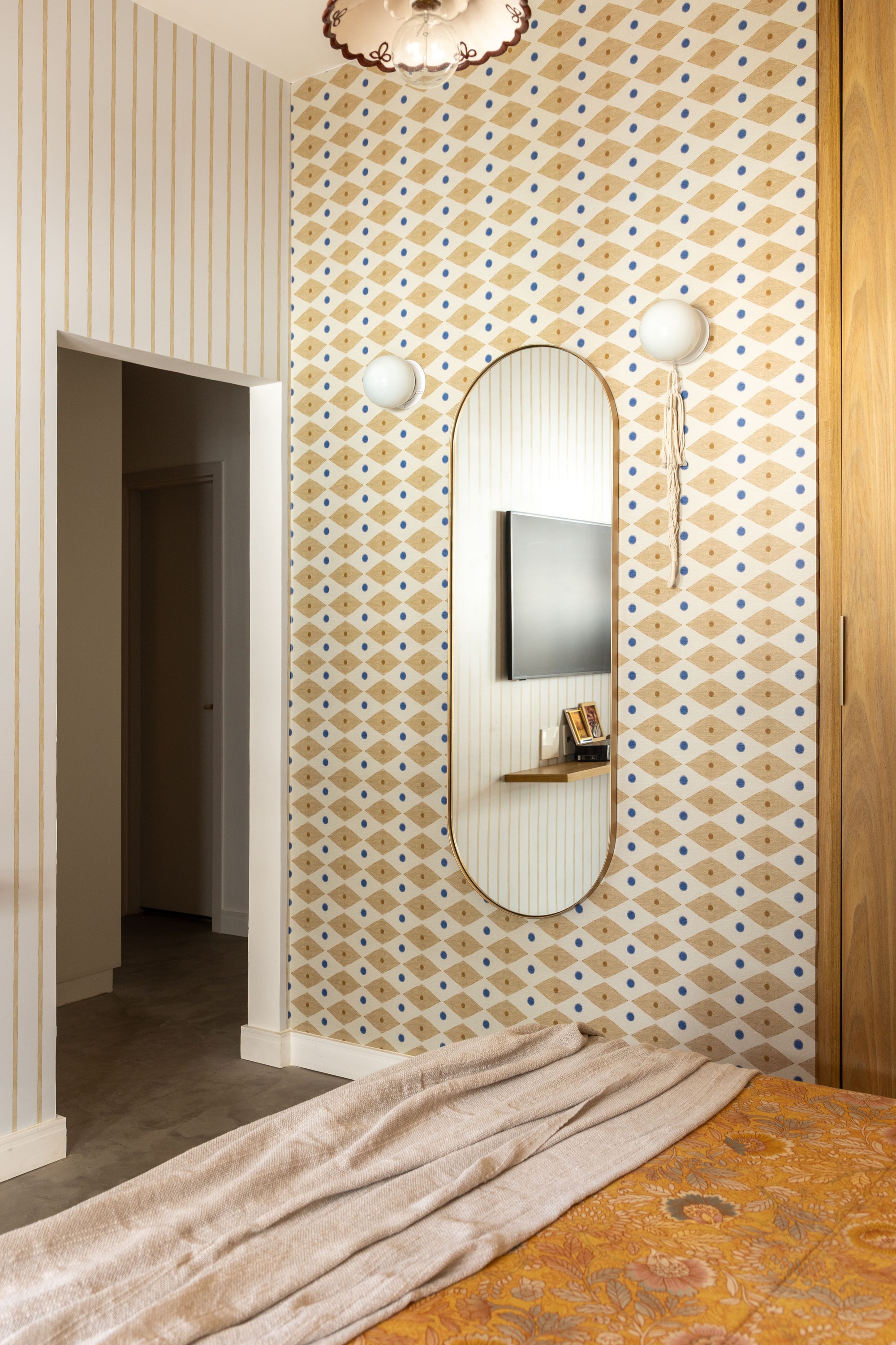 Bedroom wall covered in diamond-patterned wallpaper with an oval mirror, wall-mounted television and globe sconces beside an open doorway