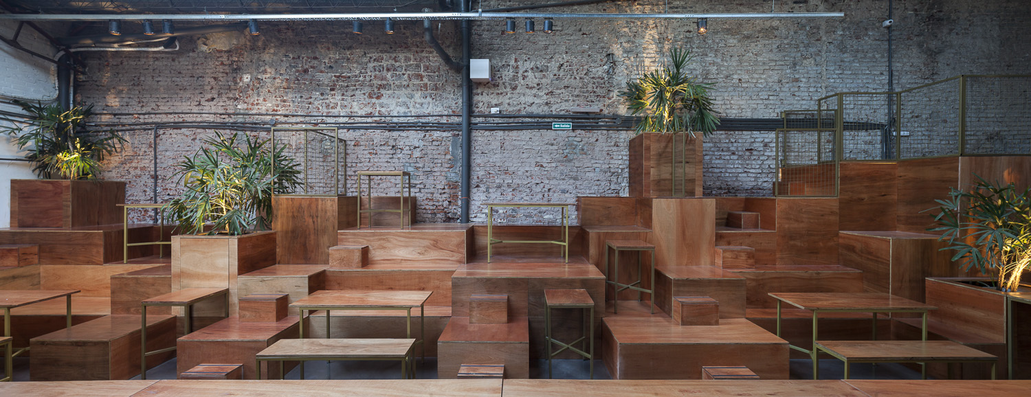 Stepped timber platforms with potted palms and brass-framed tables against an exposed brick wall under track lighting